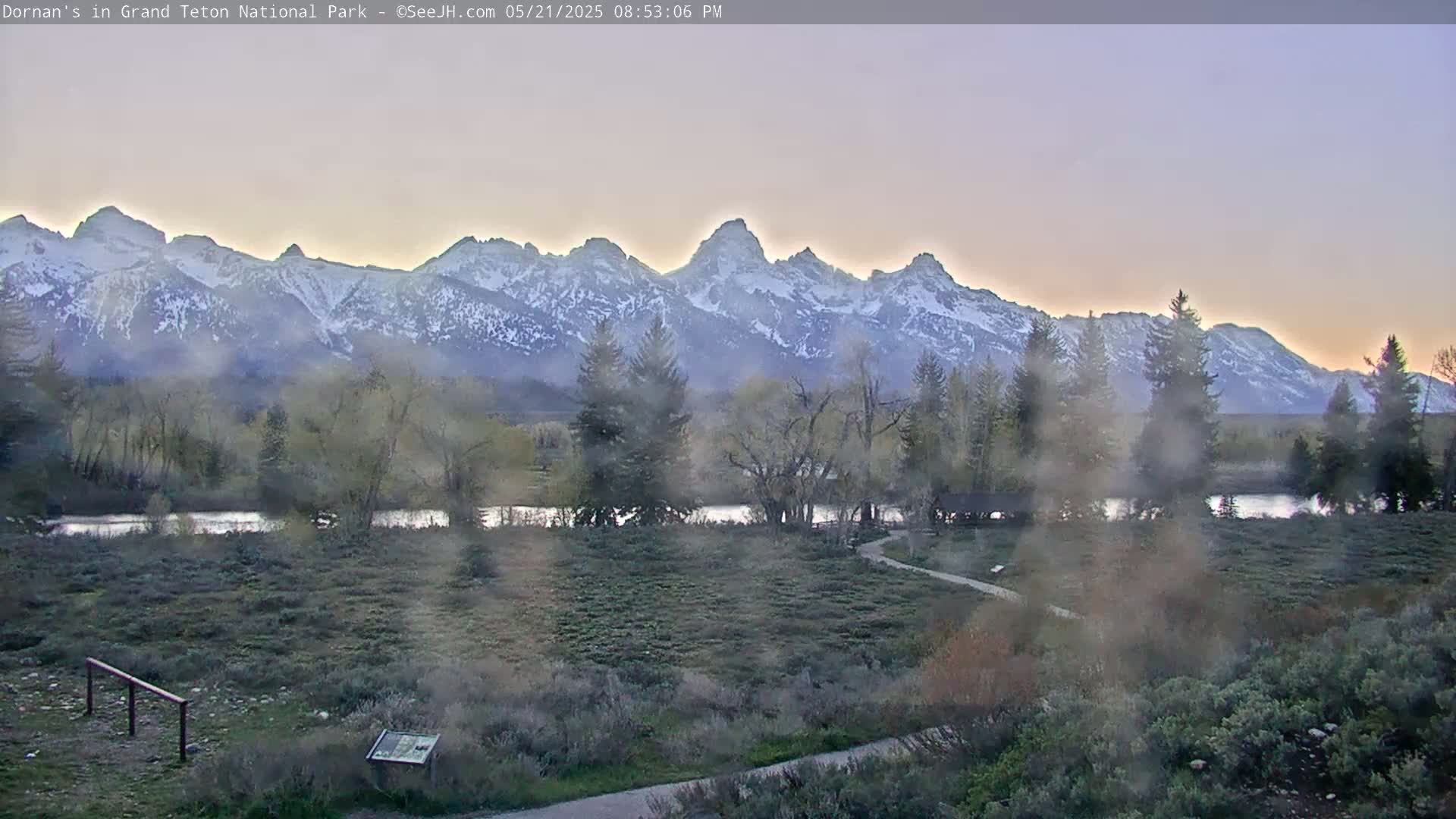 A hazy sunset illuminates snow-capped mountains behind a flat, grassy plain with trees, a river, and a walking path.
