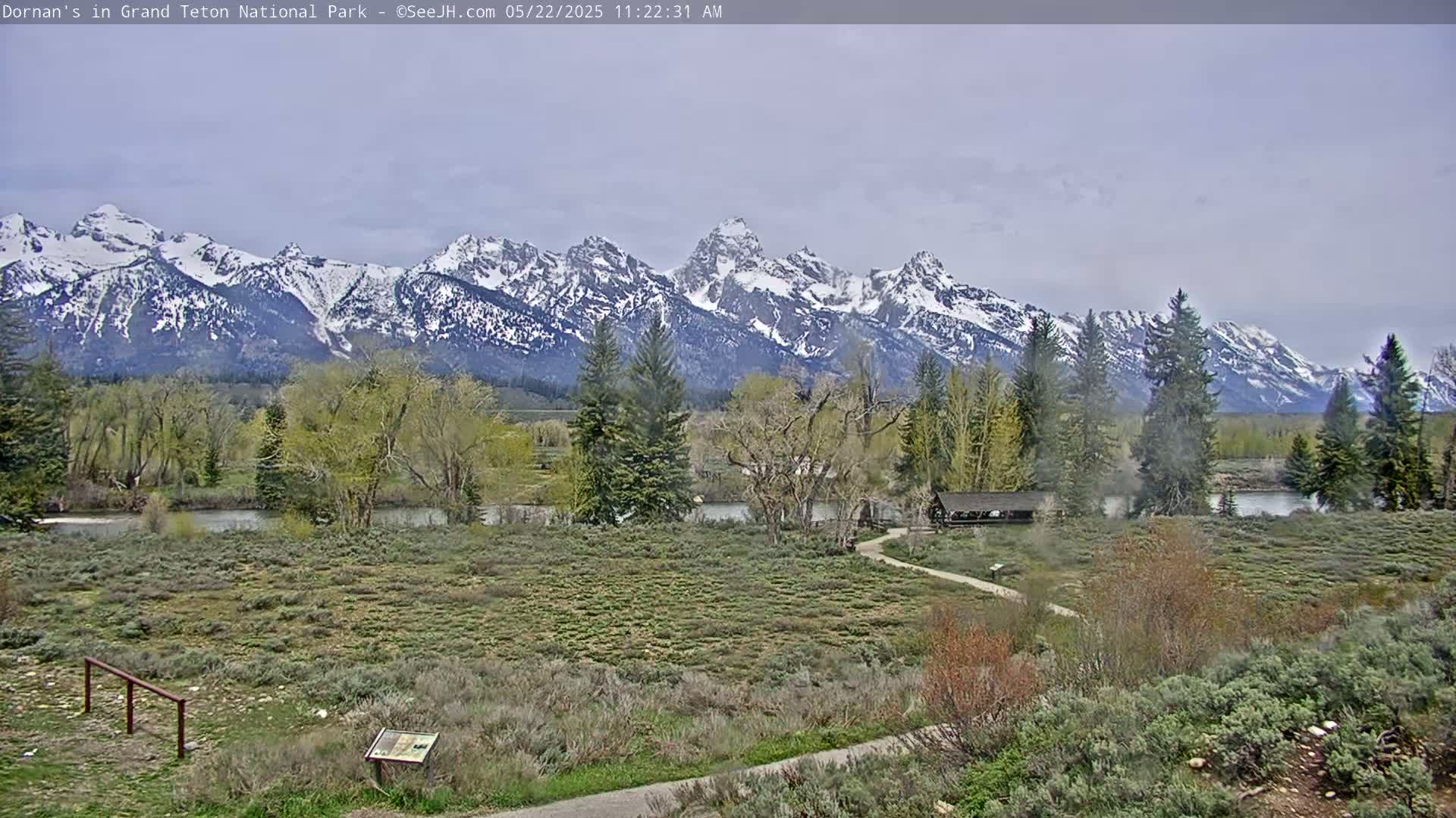 A snow-capped mountain range is visible in the background, with a flat, grassy foreground featuring trees, shrubs, a river, and a small covered structure; the sky is overcast.
