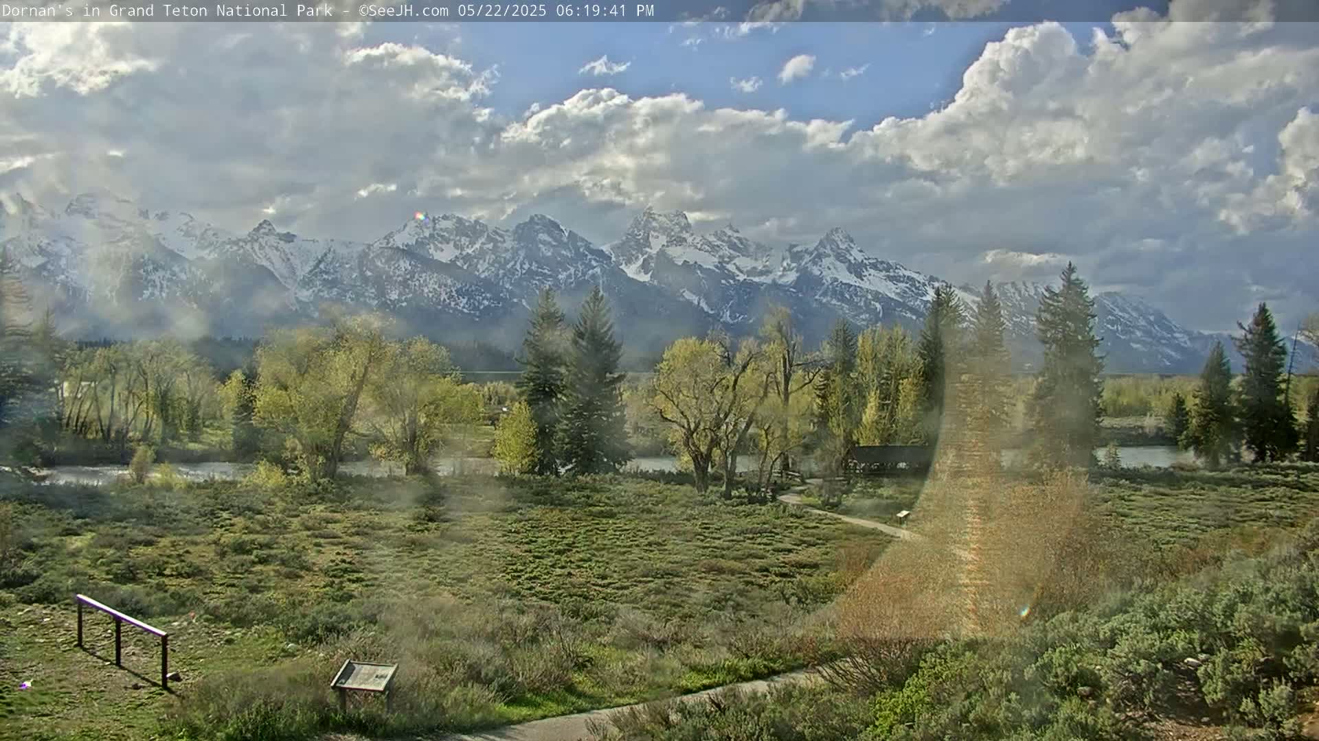 A partly cloudy day reveals a snow-capped mountain range in the distance, overlooking a flat, grassy plain dotted with trees and a small river.