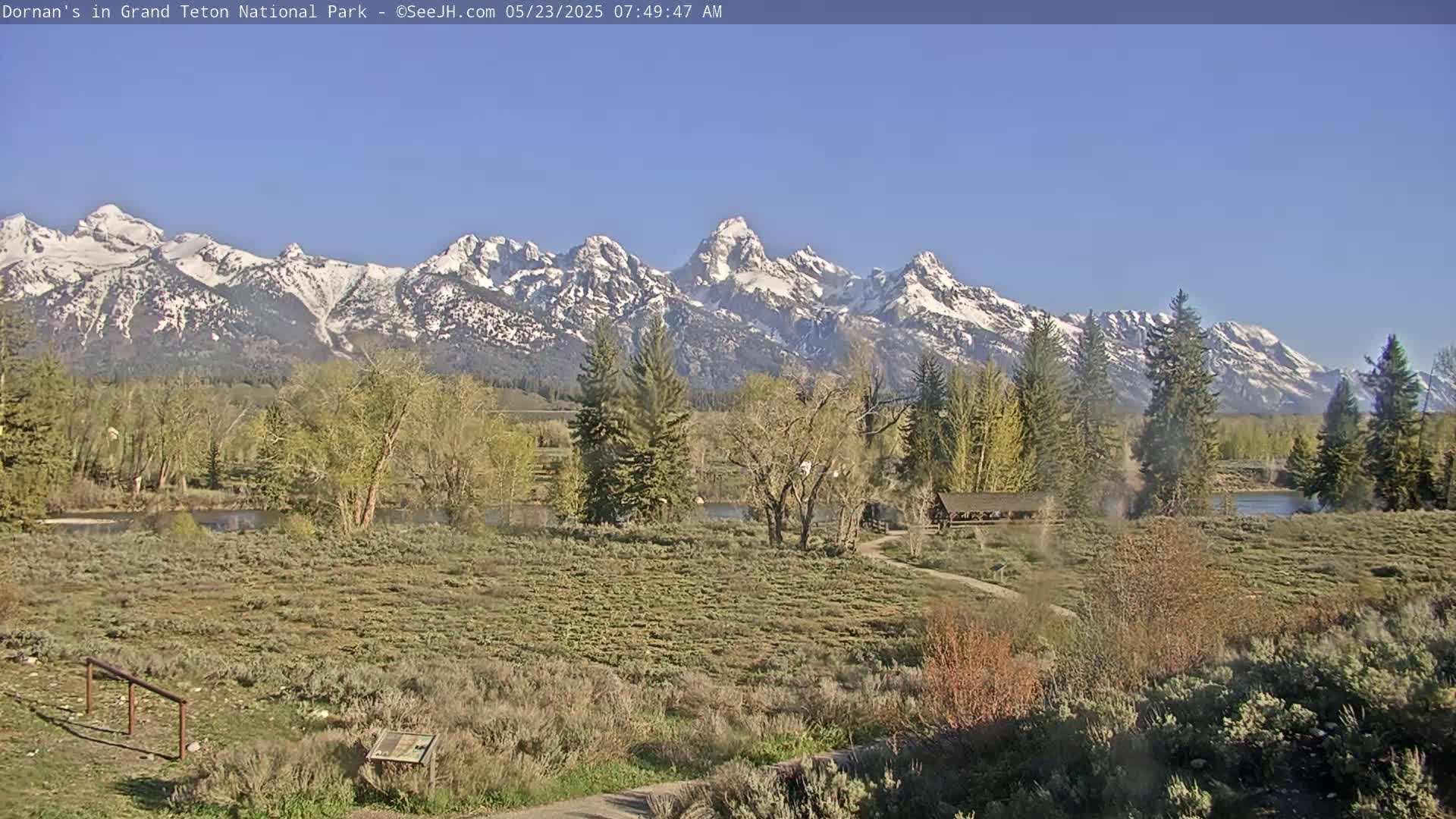 A snow-capped mountain range rises in the background of a sunny, flat landscape with a river, trees beginning to bud, and a small building.