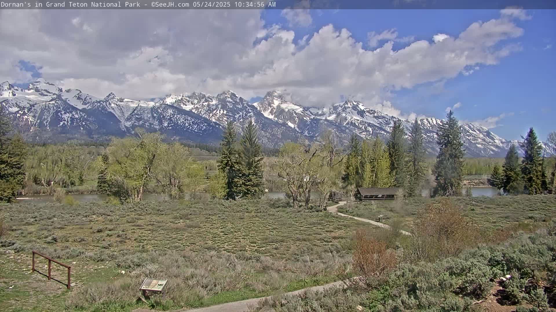 A partly cloudy day shows a view of a snow-capped mountain range behind a flat, grassy area with trees and a small building near a river.