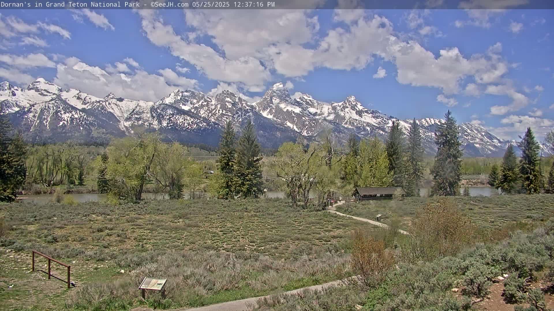 A partly cloudy day shows a snow-capped mountain range in the background, with a flat foreground of green shrubs, trees, and a calm river, all in front of a small wooden structure.