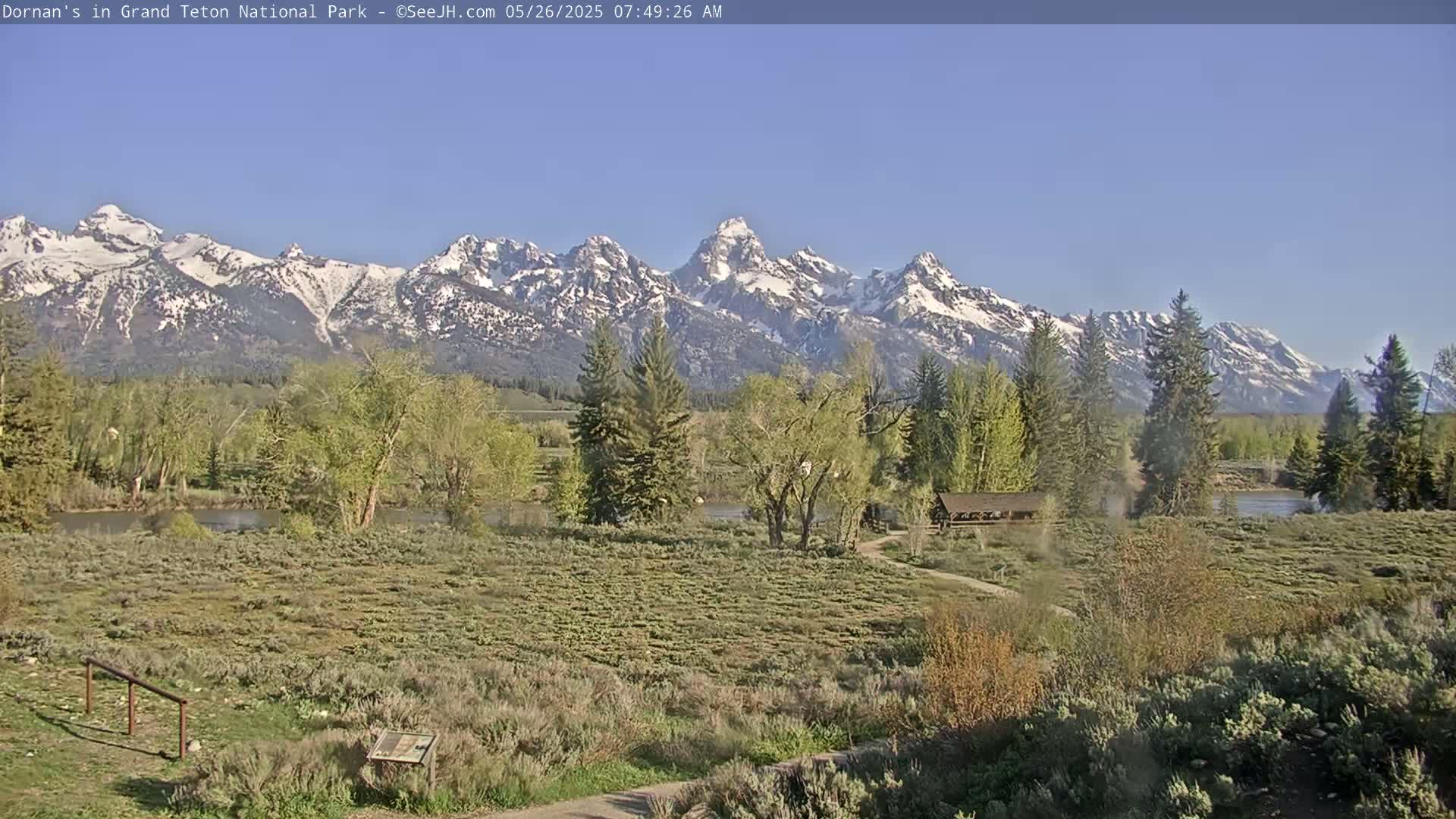 A snow-capped mountain range is visible in the background of a sunny landscape featuring a river, trees, shrubbery, and a small building.