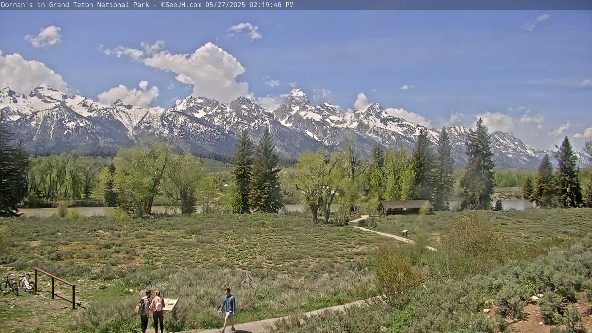 A partly cloudy day shows snow-capped mountains in the background, with a flat, grassy foreground featuring a few people walking on a path near a river and some trees.