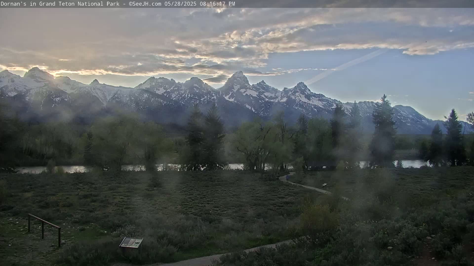 A hazy, partly cloudy evening scene shows a river winding through a flat, shrubby area before a range of snow-capped mountains.