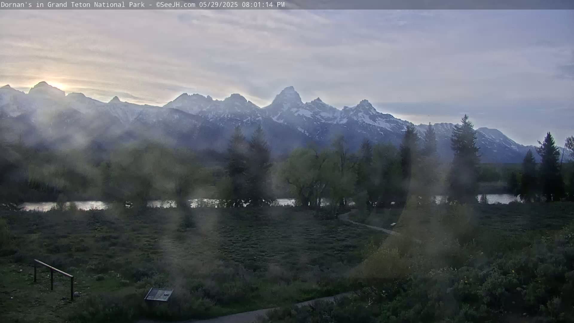 A hazy, late-afternoon view of snow-capped mountains across a flat, grassy plain with a river and some trees, under a partly cloudy sky.
