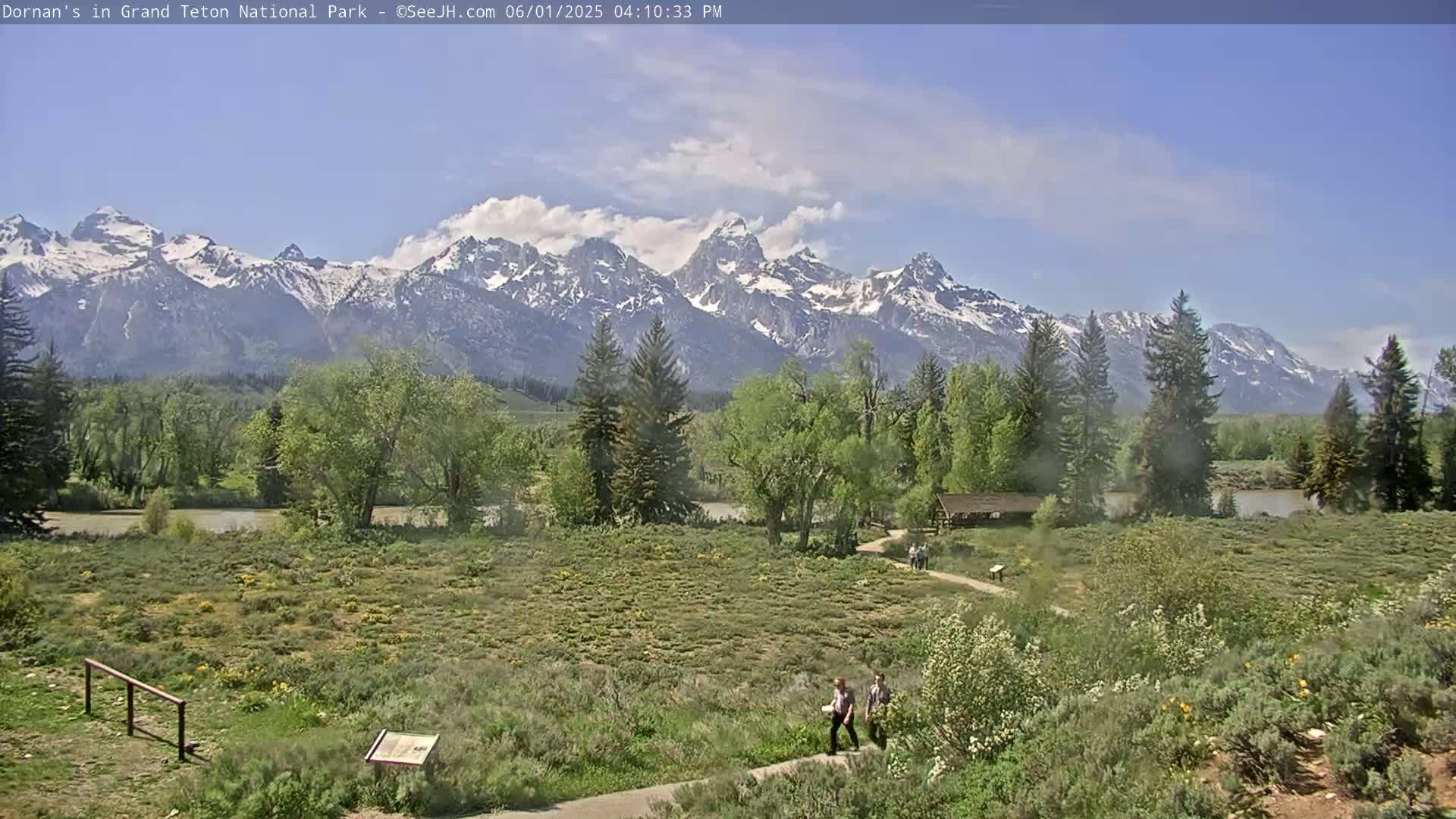 A partly cloudy day reveals a snow-capped mountain range in the background, overlooking a grassy field with two people walking on a path near trees and a calm river.