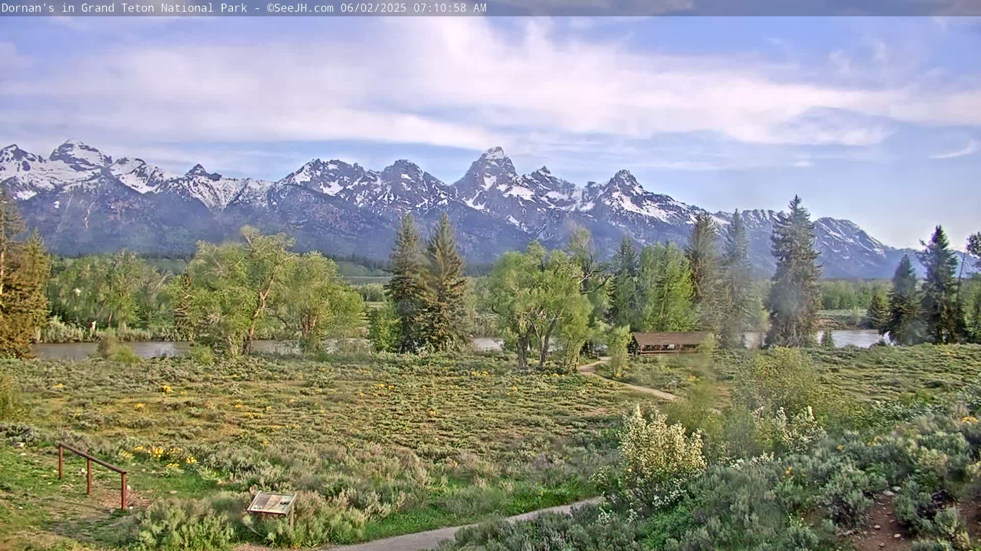 A partly cloudy day reveals a snow-capped mountain range behind a flat, green field with a river and a few trees, near a small building.