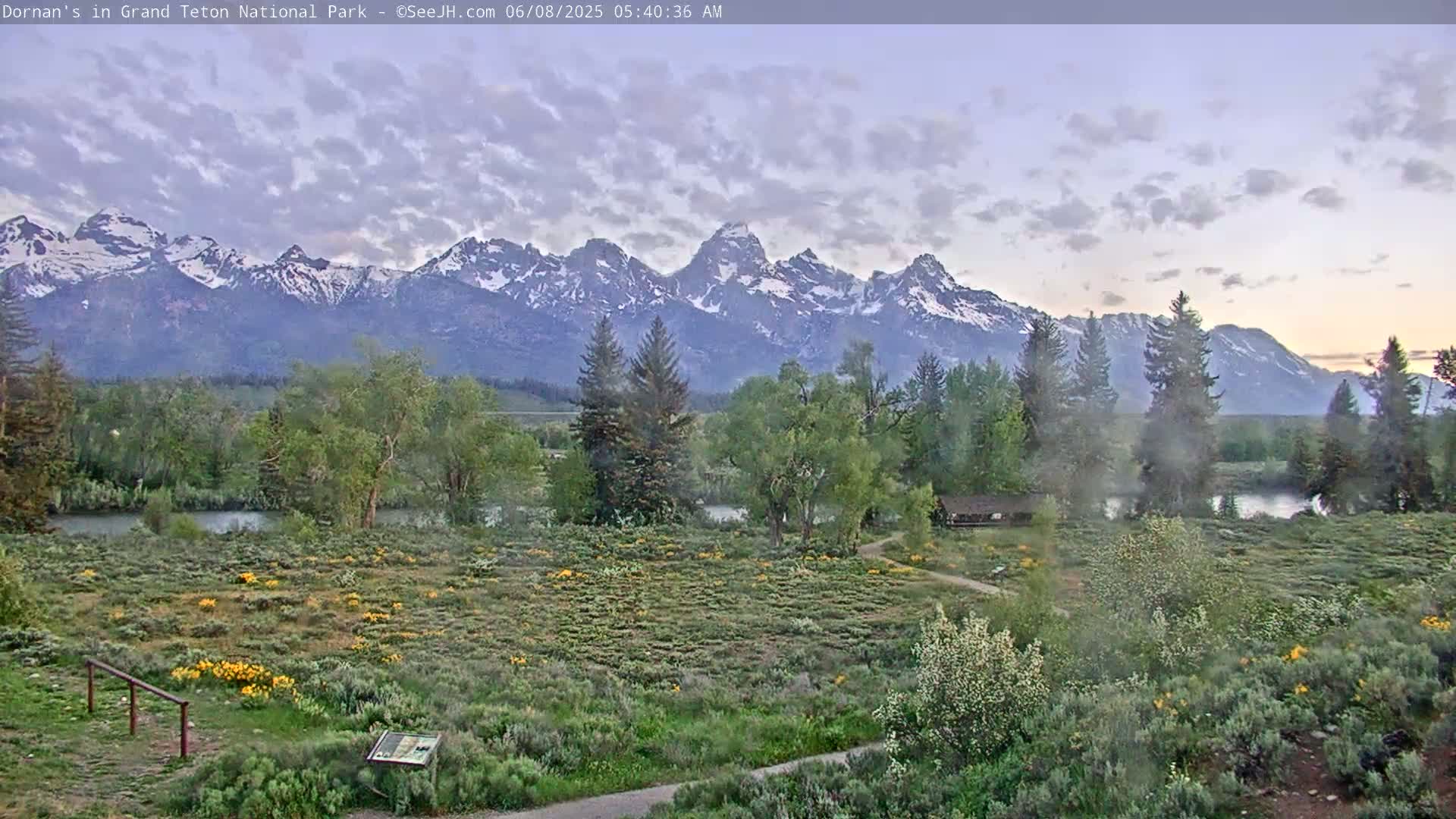 A partly cloudy dawn reveals a snow-capped mountain range in the distance, overlooking a flat, grassy field with trees and a small building near a calm river.