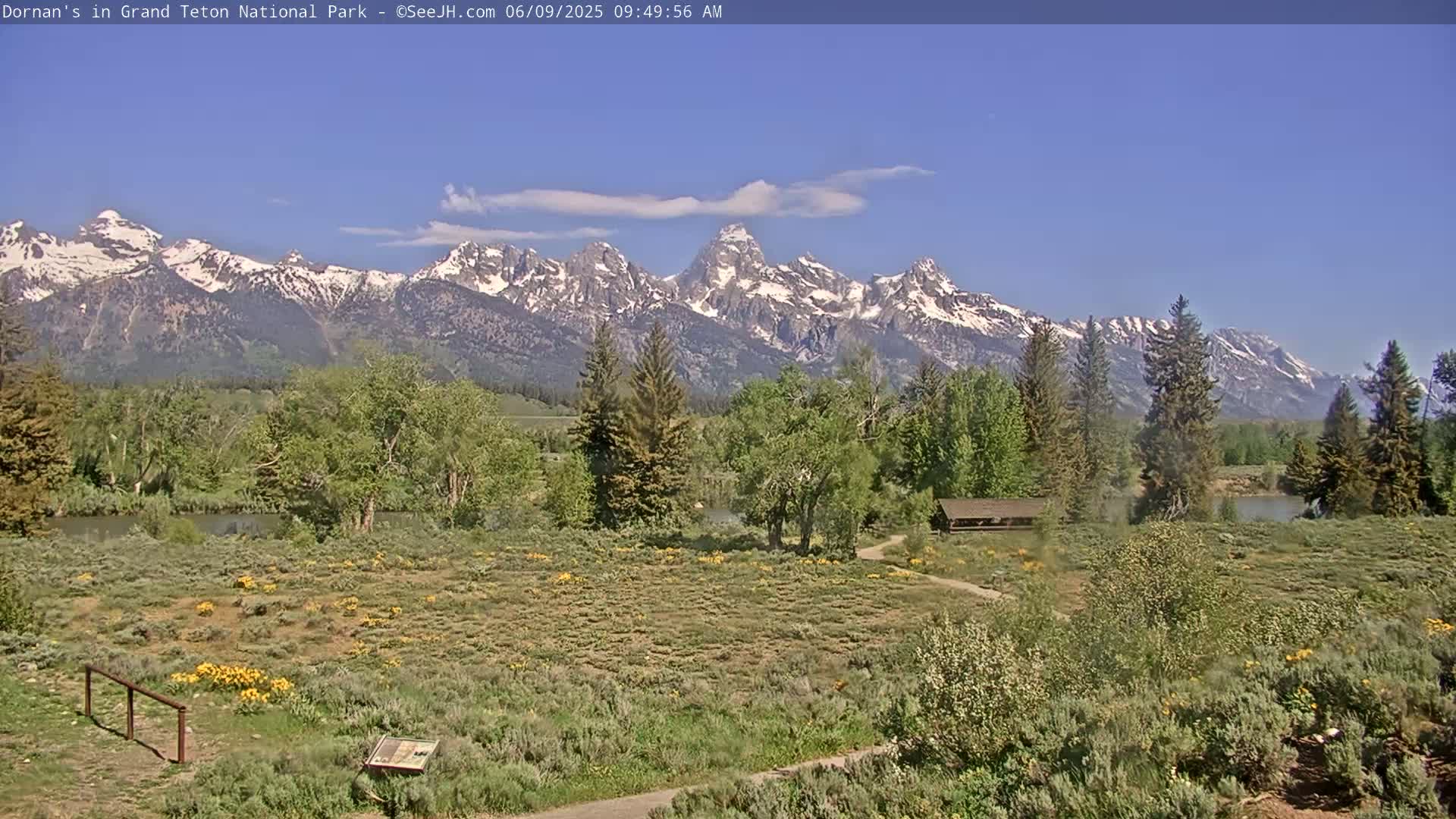 A picturesque landscape shows a grassy field with a small pavilion, bordered by trees and shrubs, with a snow-capped mountain range in the background under a mostly clear, blue sky.