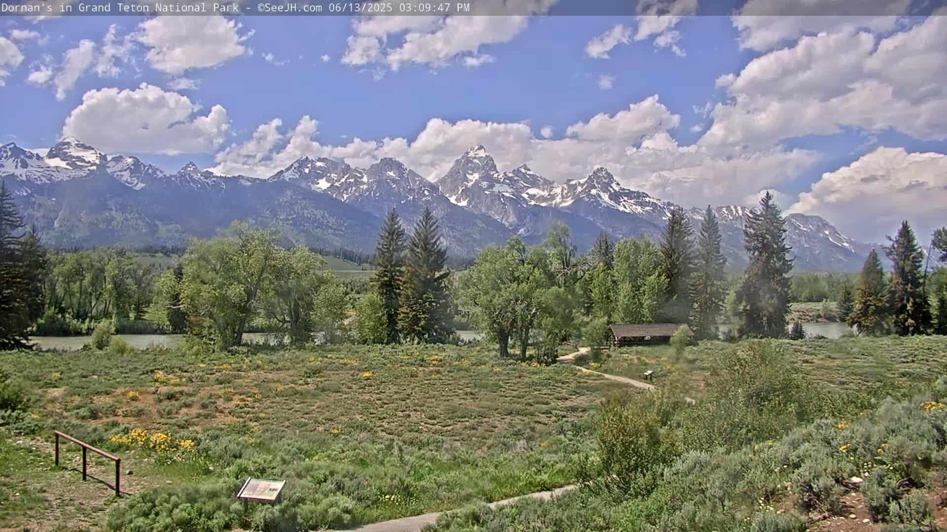 A partly cloudy sky overlooks a verdant valley with a river, trees, and a snow-capped mountain range in the distance.