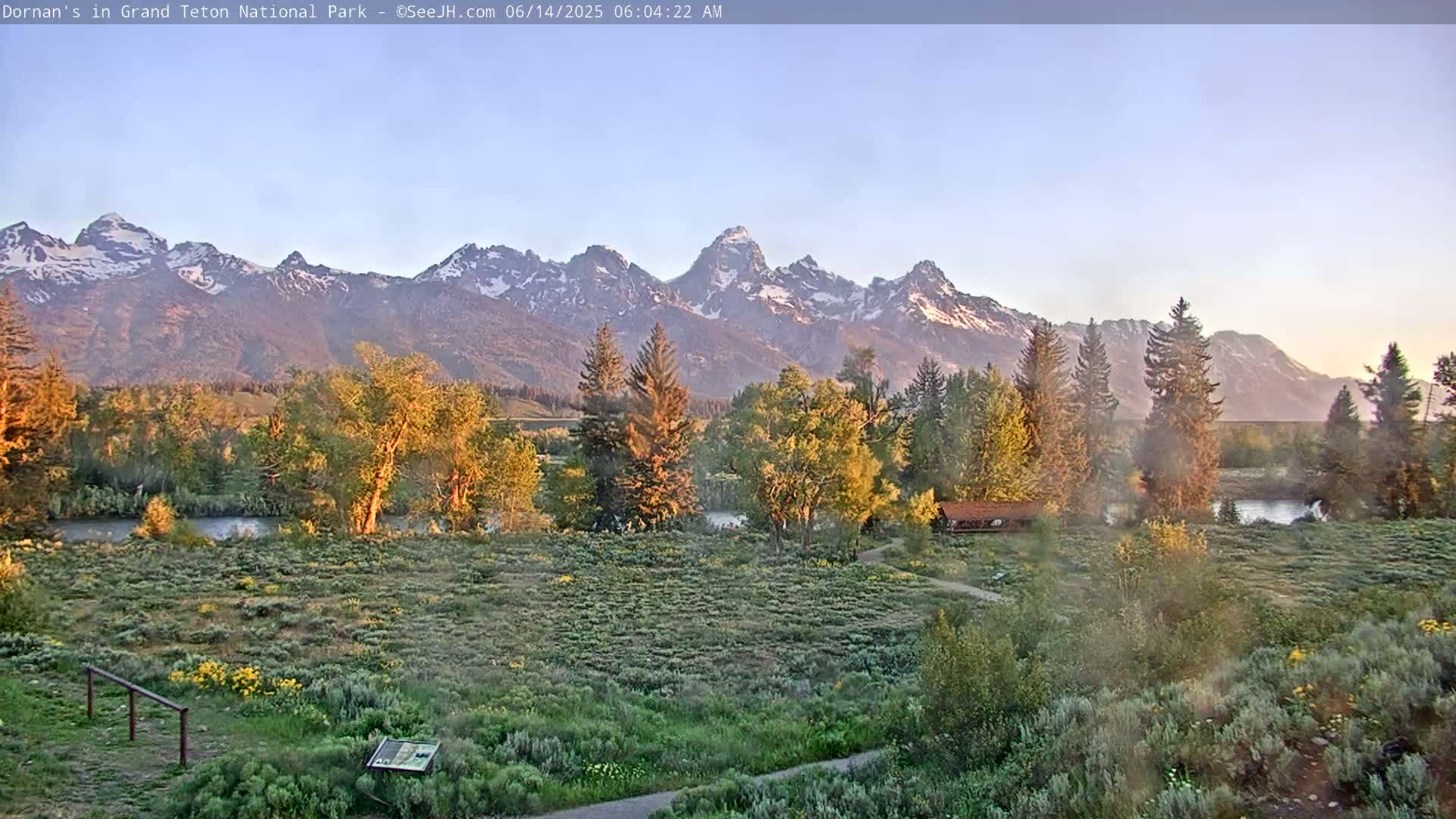 A clear morning reveals a picturesque landscape of a grassy field, trees, a river, and a snow-capped mountain range in the distance.
