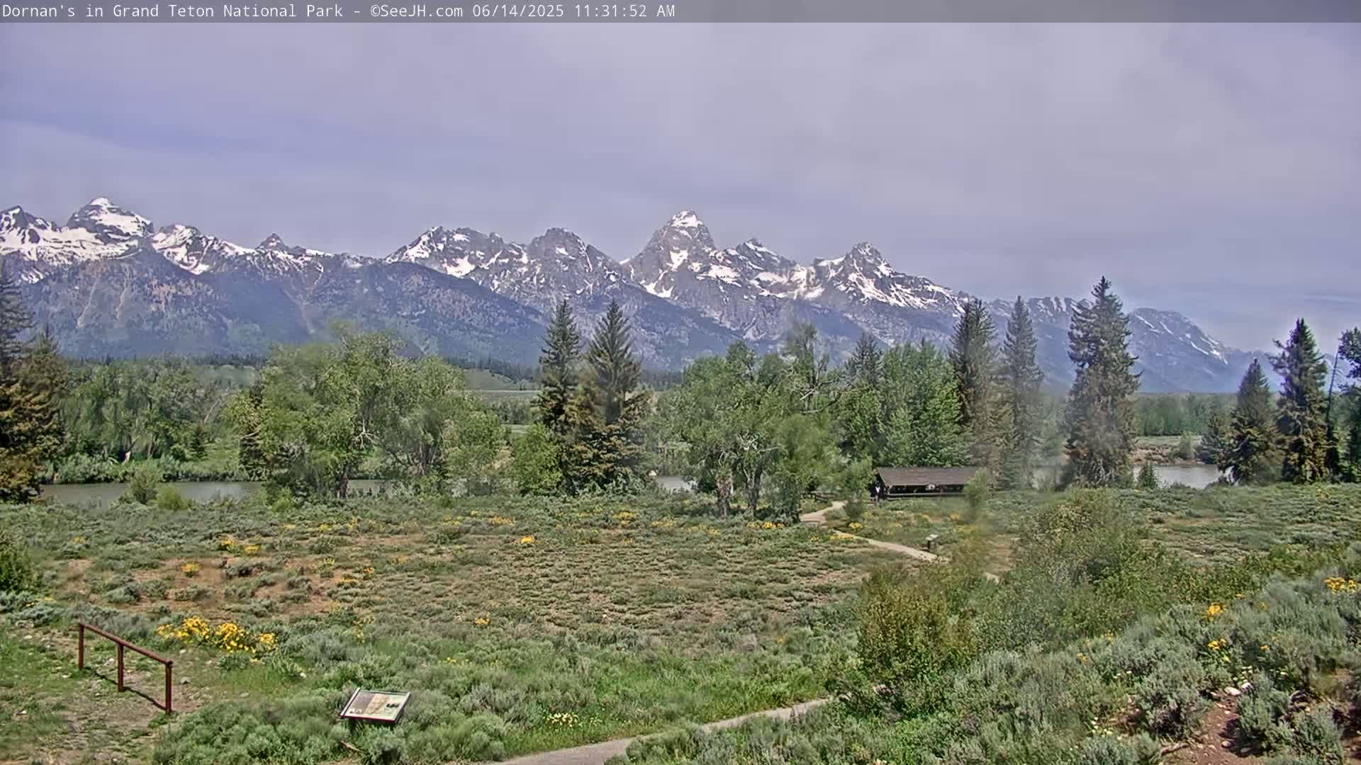 A partly cloudy day reveals a tranquil river scene with lush green vegetation, a covered pavilion, and snow-capped mountains in the background.
