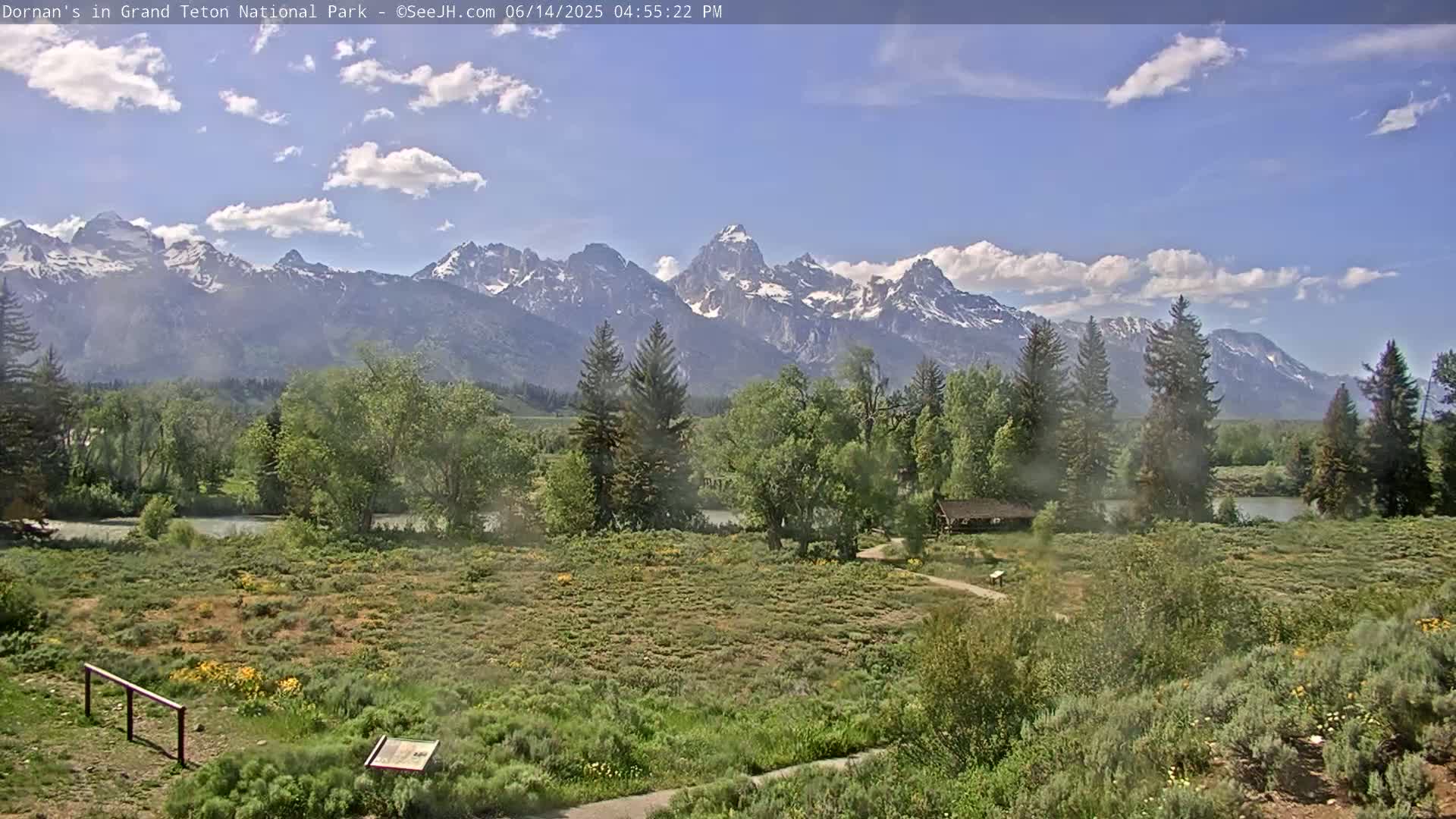 A partly cloudy day reveals a tranquil river scene with lush green vegetation, a covered pavilion, and snow-capped mountains in the background.