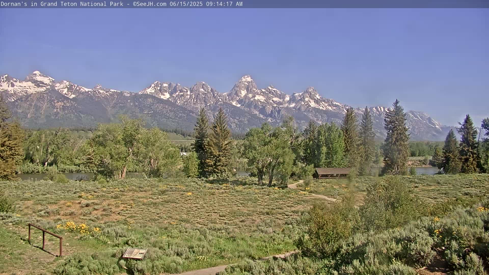 A sunny day reveals a tranquil scene of a flat, grassy field with a few trees and a small shelter, overlooking a river and a snow-capped mountain range in the background.