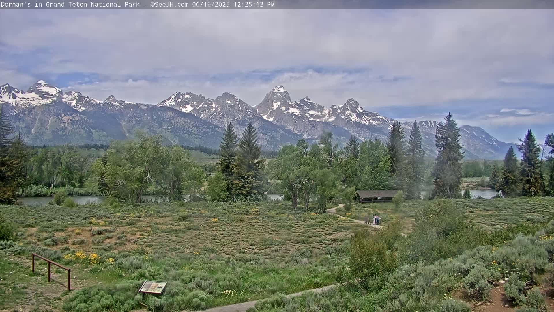 A partly cloudy day reveals a snow-capped mountain range in the distance, behind a flat, grassy field with a few trees and people near a small building, and a river running through the foreground.