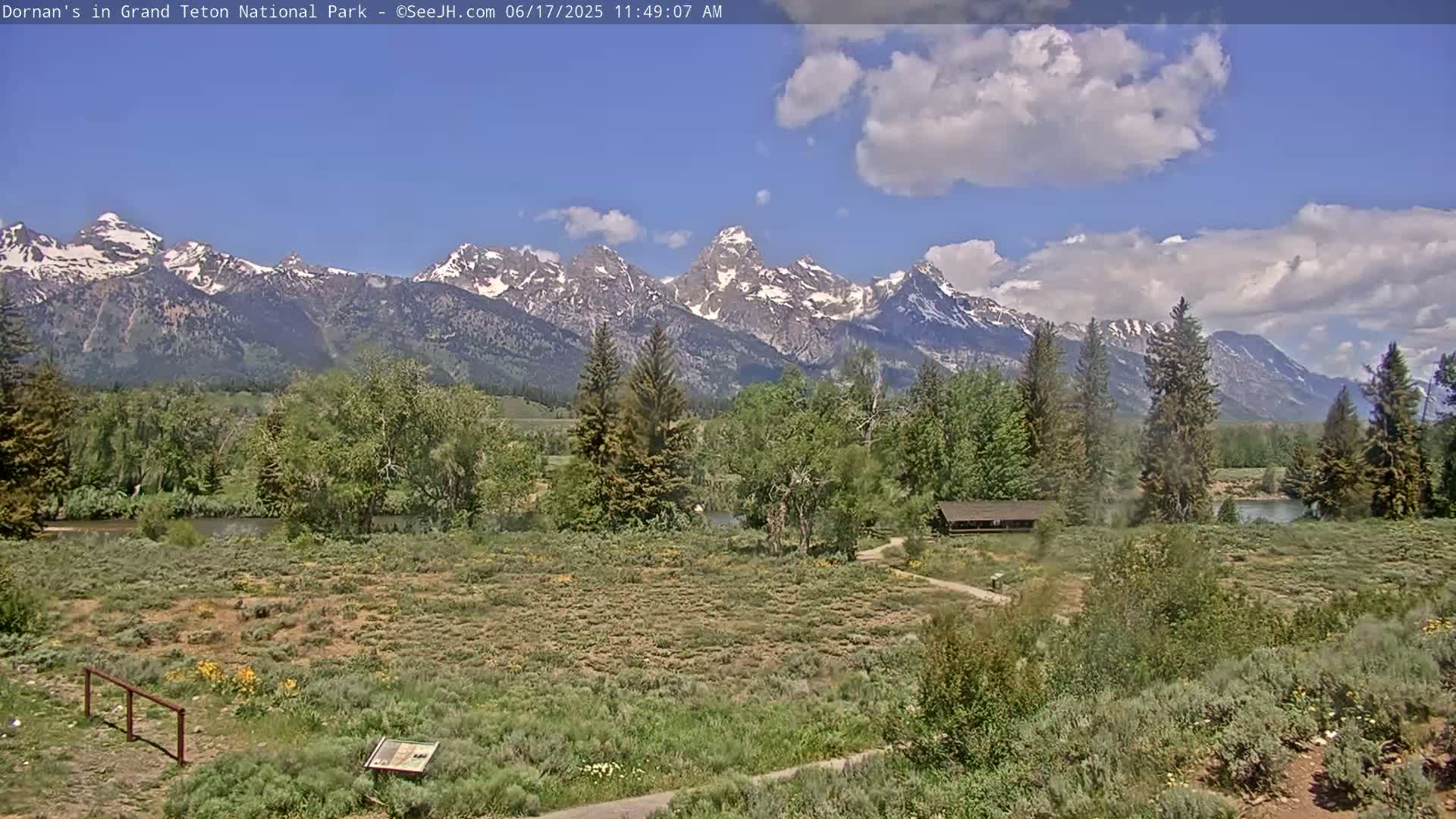 A partly cloudy day reveals a view of a river, green vegetation, a small building, and a mountain range capped with snow.