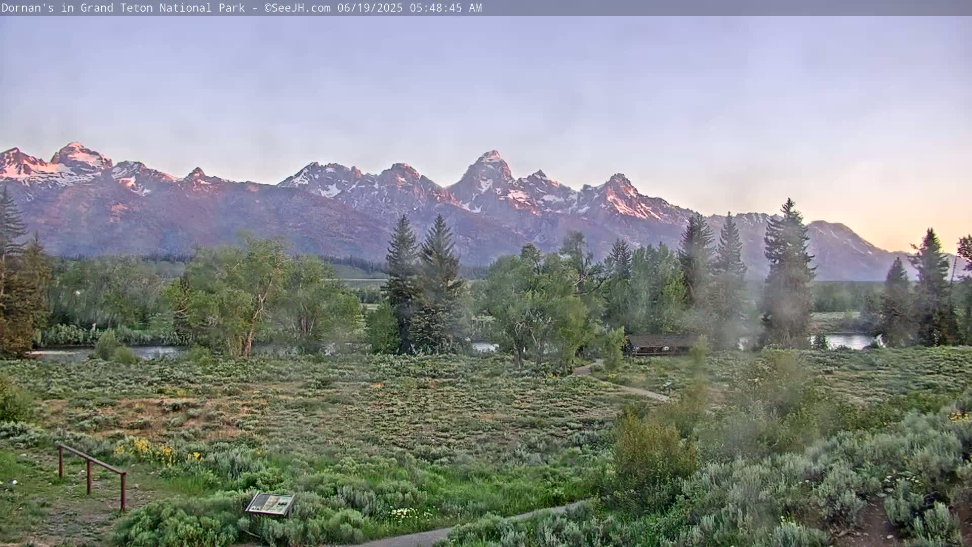 A calm sunrise illuminates a snow-capped mountain range behind a verdant valley with a river, trees, and shrubs.