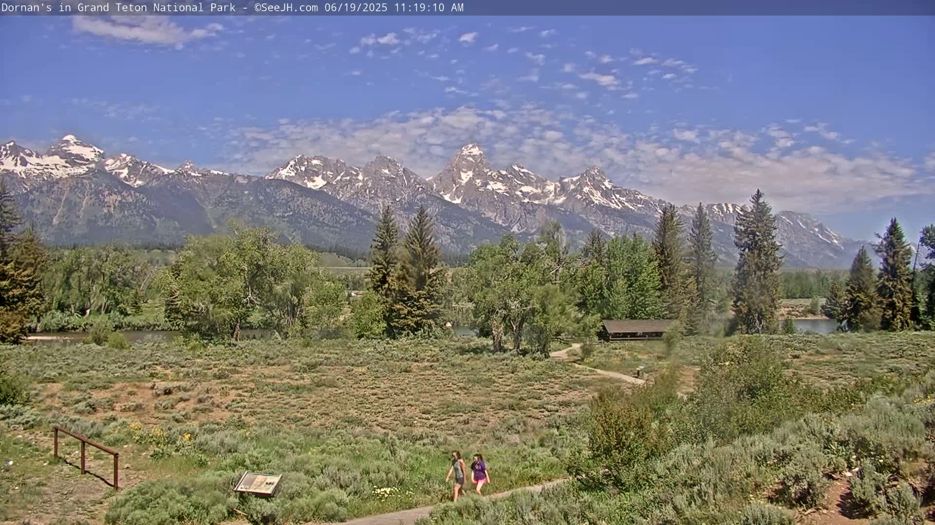Two people walk on a path through a grassy area near a river, with a mountain range in the background under a mostly sunny sky.
