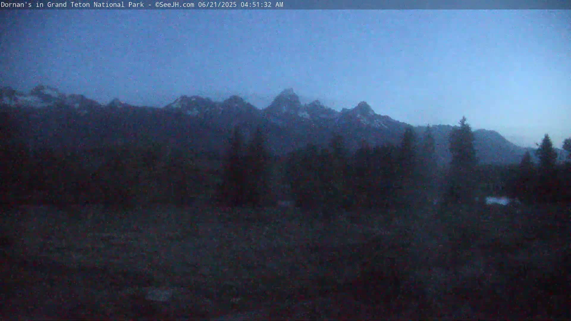 A dark, pre-dawn view shows a mountain range in the distance, silhouetted against a twilight sky, with a foreground of dark trees and brush.