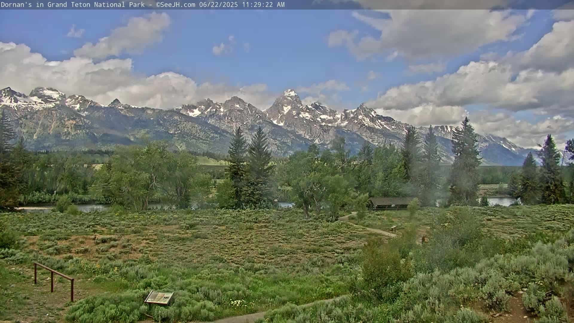 A partly cloudy day reveals a picturesque valley with a river, lush greenery, and snow-capped mountains in the background.