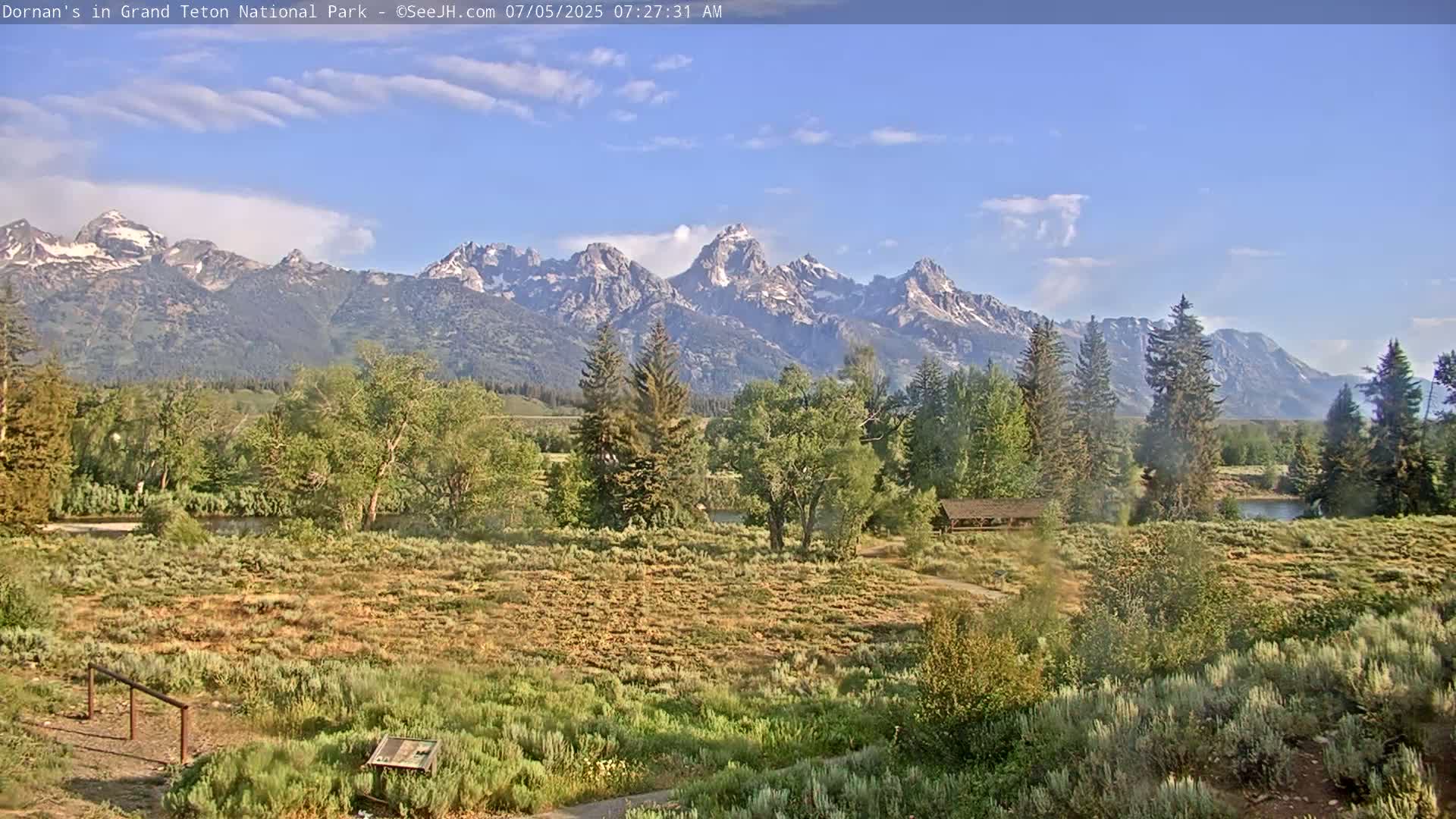 A partly sunny day reveals a field of low-lying vegetation and trees in front of a majestic mountain range under a mostly clear sky.