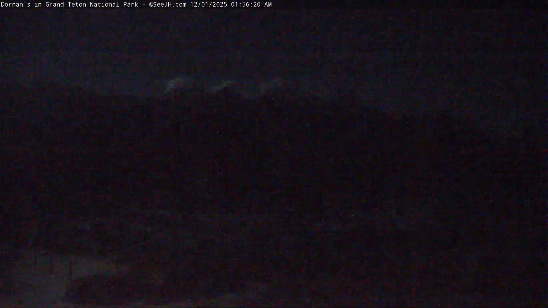 An extremely dark, grainy nighttime scene reveals faint, hazy horizontal shapes across the upper middle, likely illuminated clouds or distant mountain peaks, under a dark and partially cloudy sky.