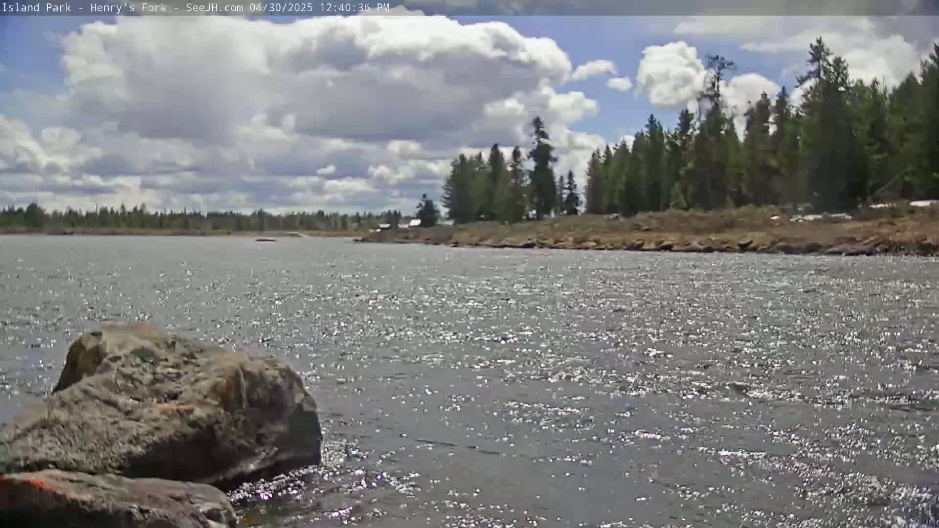 A partly cloudy day reveals a wide river flowing past a rocky shoreline and a background of evergreen trees.
