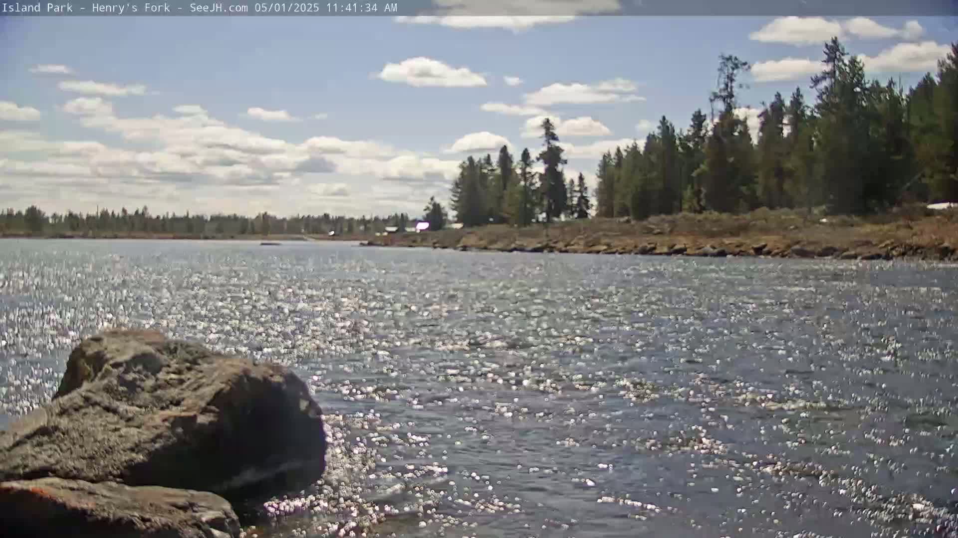 A rocky shoreline borders a gently flowing river under a partly cloudy sky.
