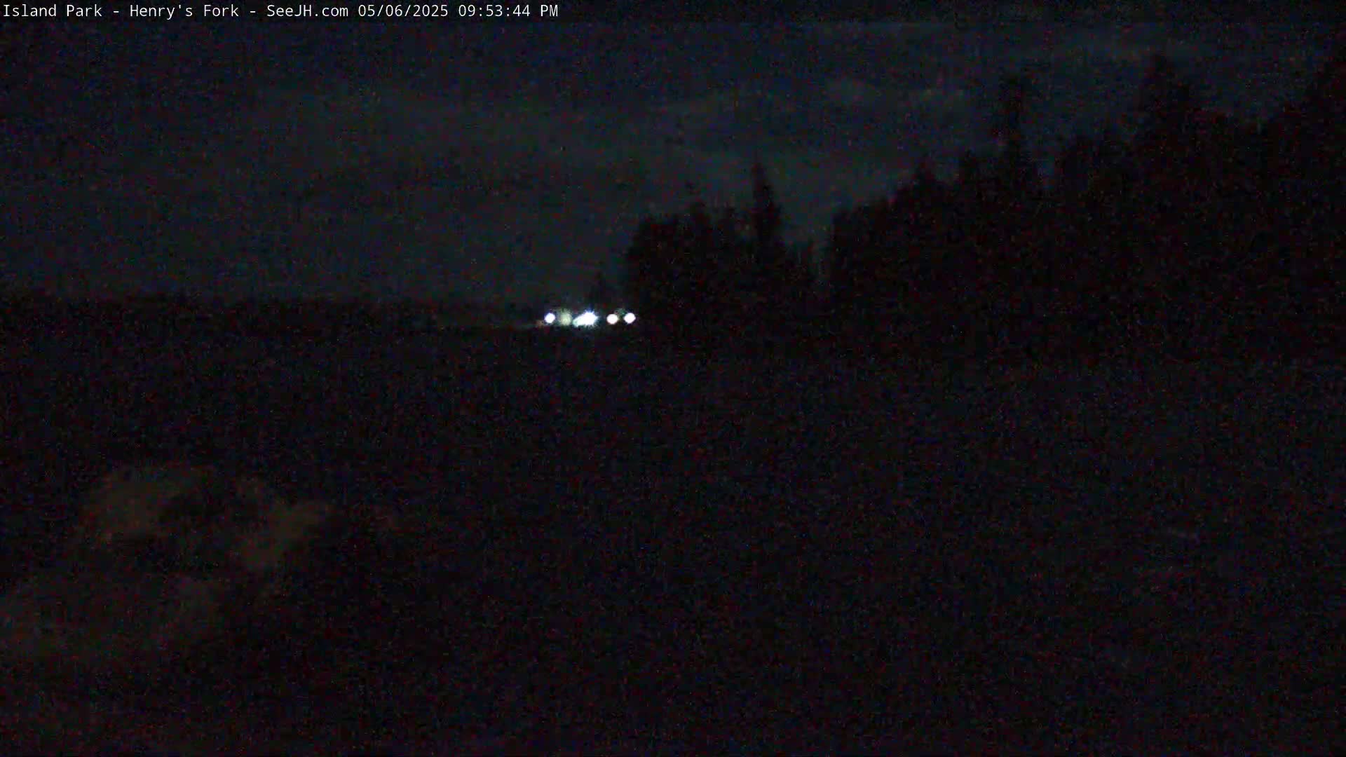 At night, the headlights of a vehicle are visible in the distance, with a dark, silhouetted treeline in the background.