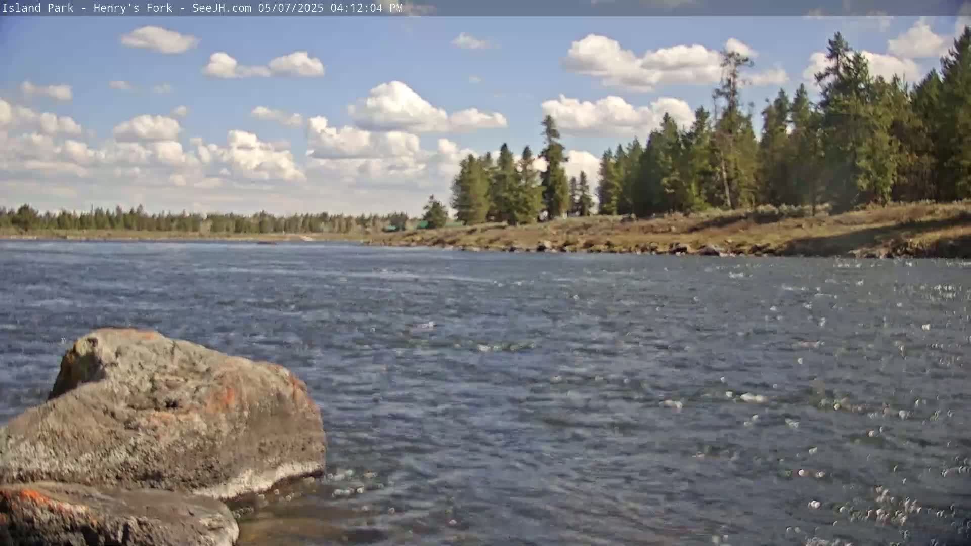 A partly cloudy sky overlooks a rocky riverbank with a flowing river and a stand of evergreen trees in the background.