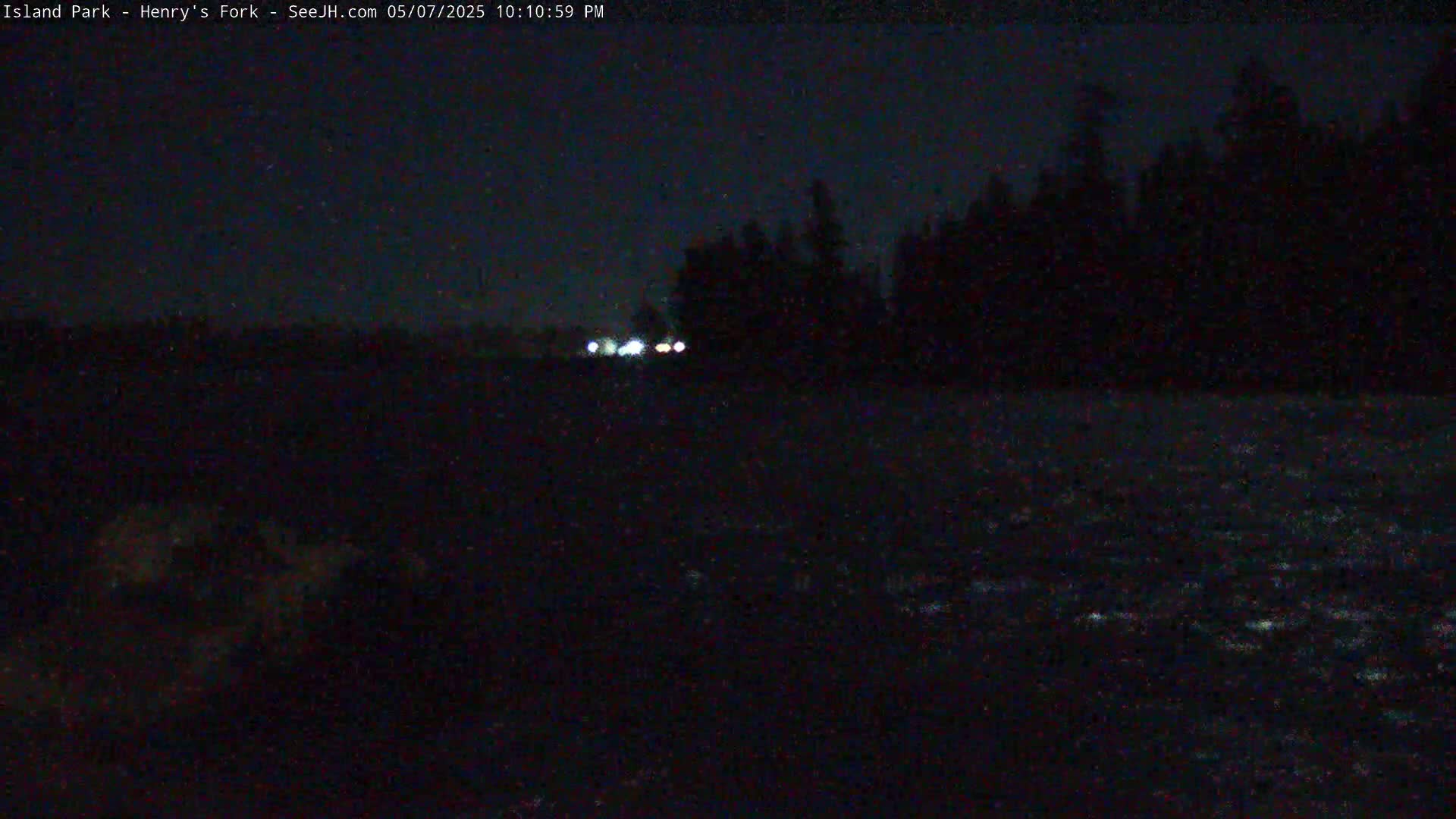 At night, headlights from a vehicle are visible in the distance across dark water, with a silhouetted treeline in the background.