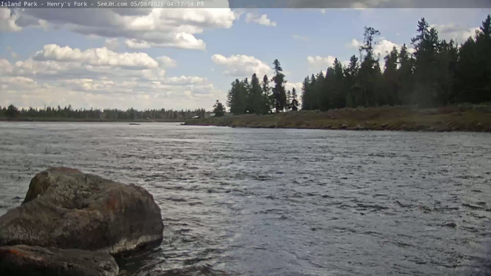 A partly cloudy sky overlooks a wide, gently flowing river with a rocky shore in the foreground and a line of dark green trees on the opposite bank.