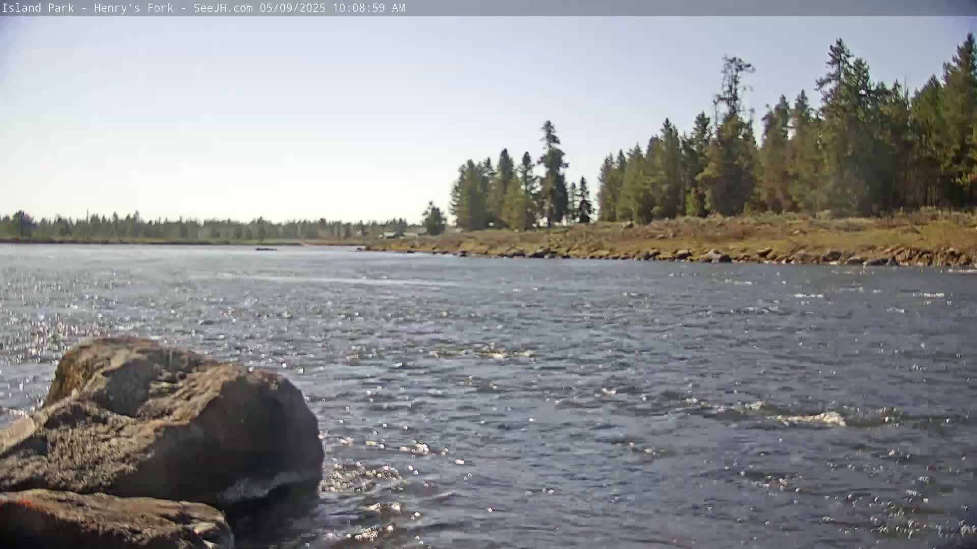 A rocky riverbank with large rocks in the foreground and a flowing river leading to a tree-lined shore under a sunny sky.