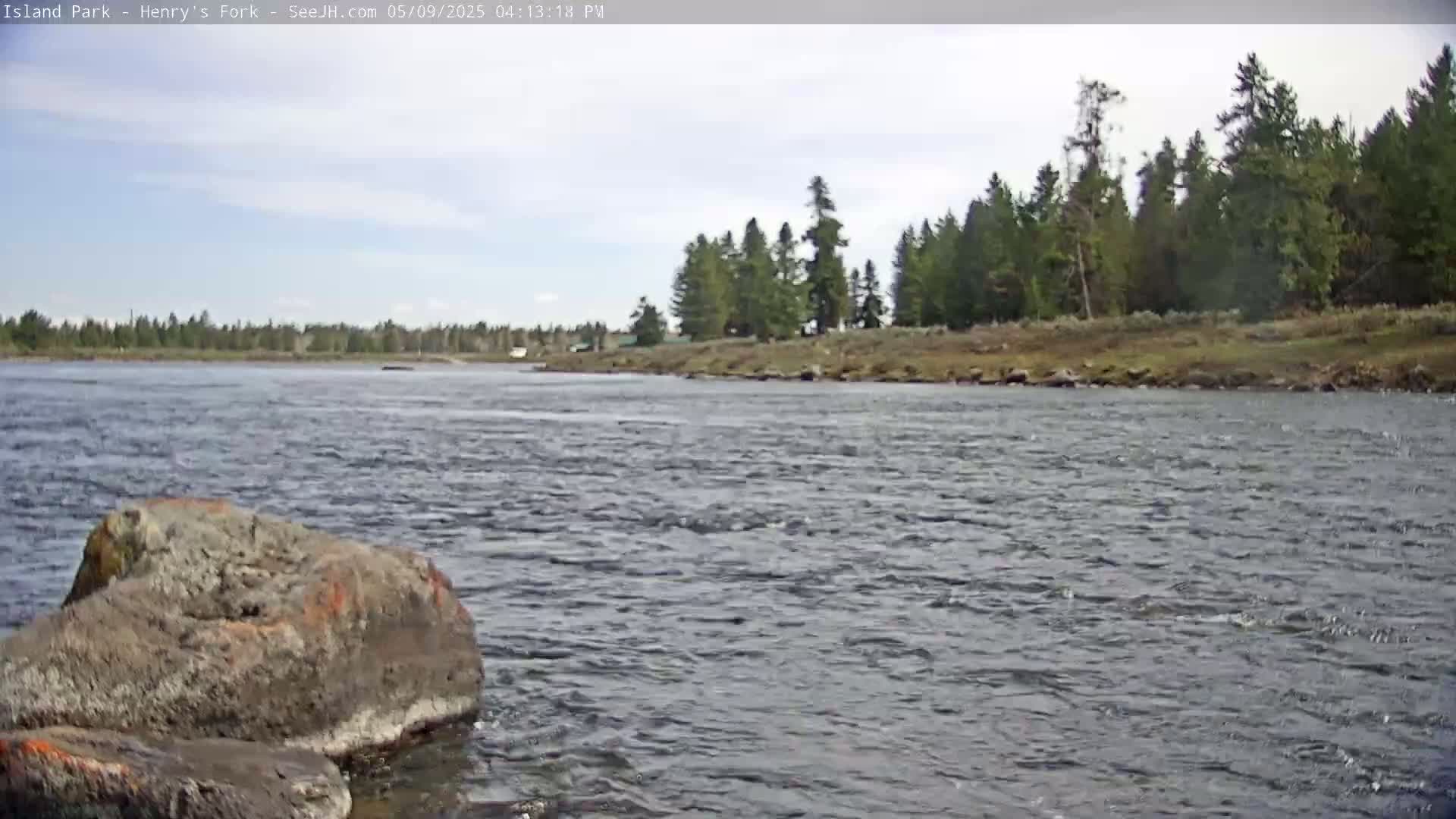 A rocky riverbank with a large gray rock in the foreground overlooks a flowing river under a partly cloudy sky, with a line of evergreen trees in the distance.