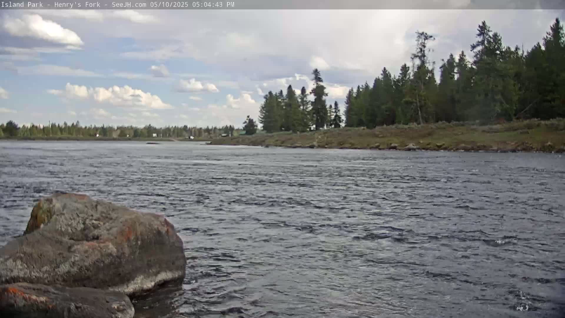 A partly cloudy sky overlooks a rocky riverbank lined with evergreen trees and a flowing river with a large rock in the foreground.