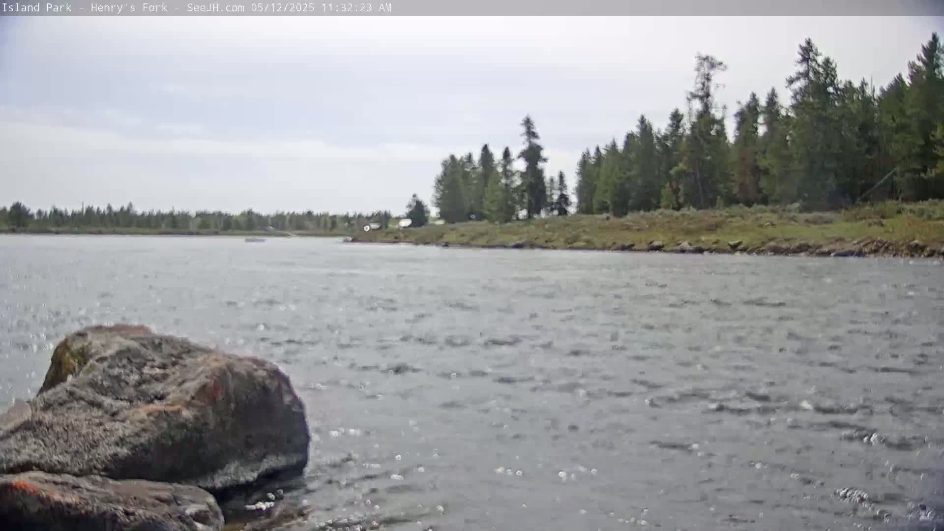 A rocky riverbank with a line of evergreens overlooks a gently flowing river under a mostly cloudy sky.