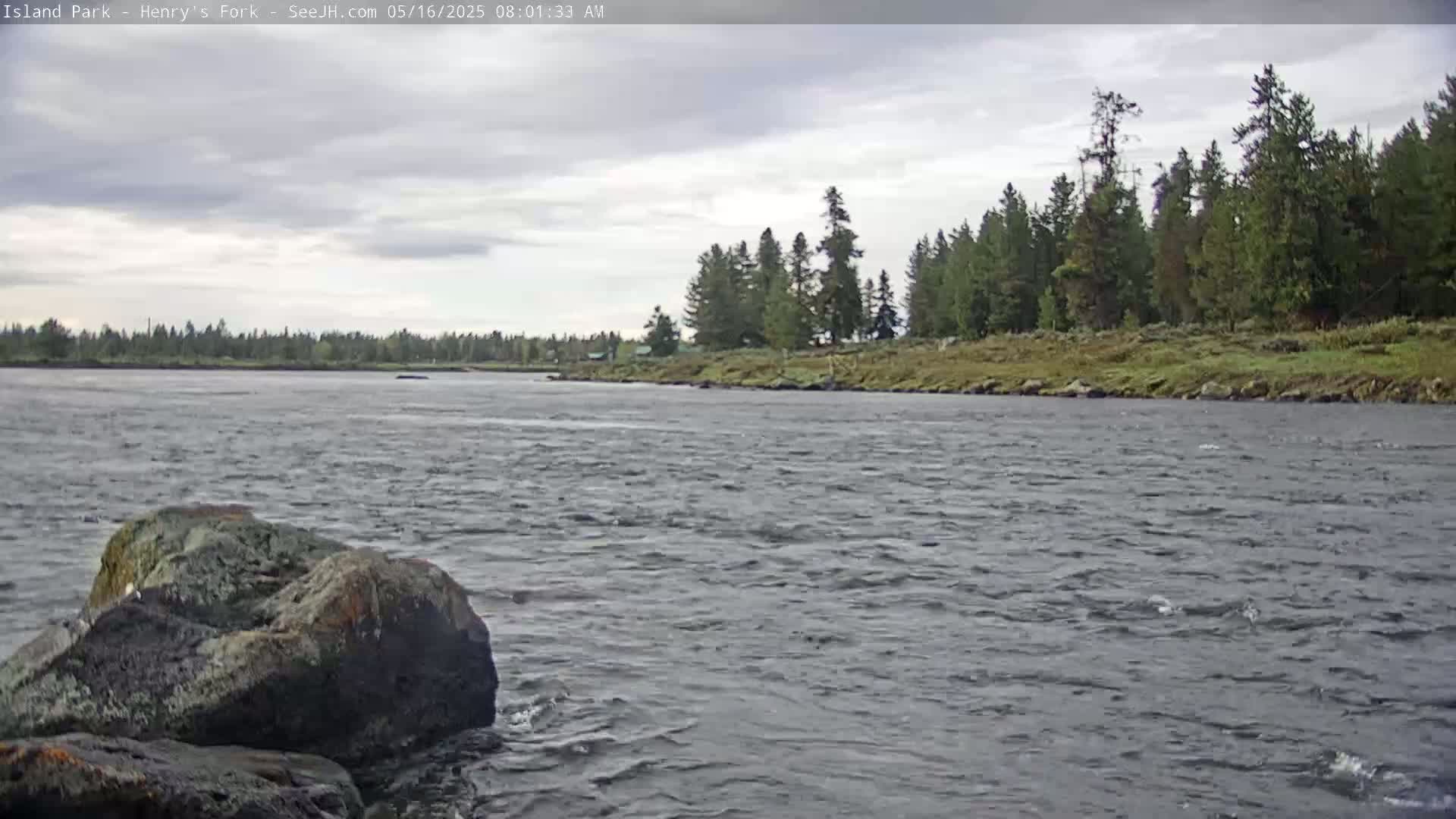 A wide, gently flowing river under a cloudy sky is bordered by a treeline and large rocks in the foreground.
