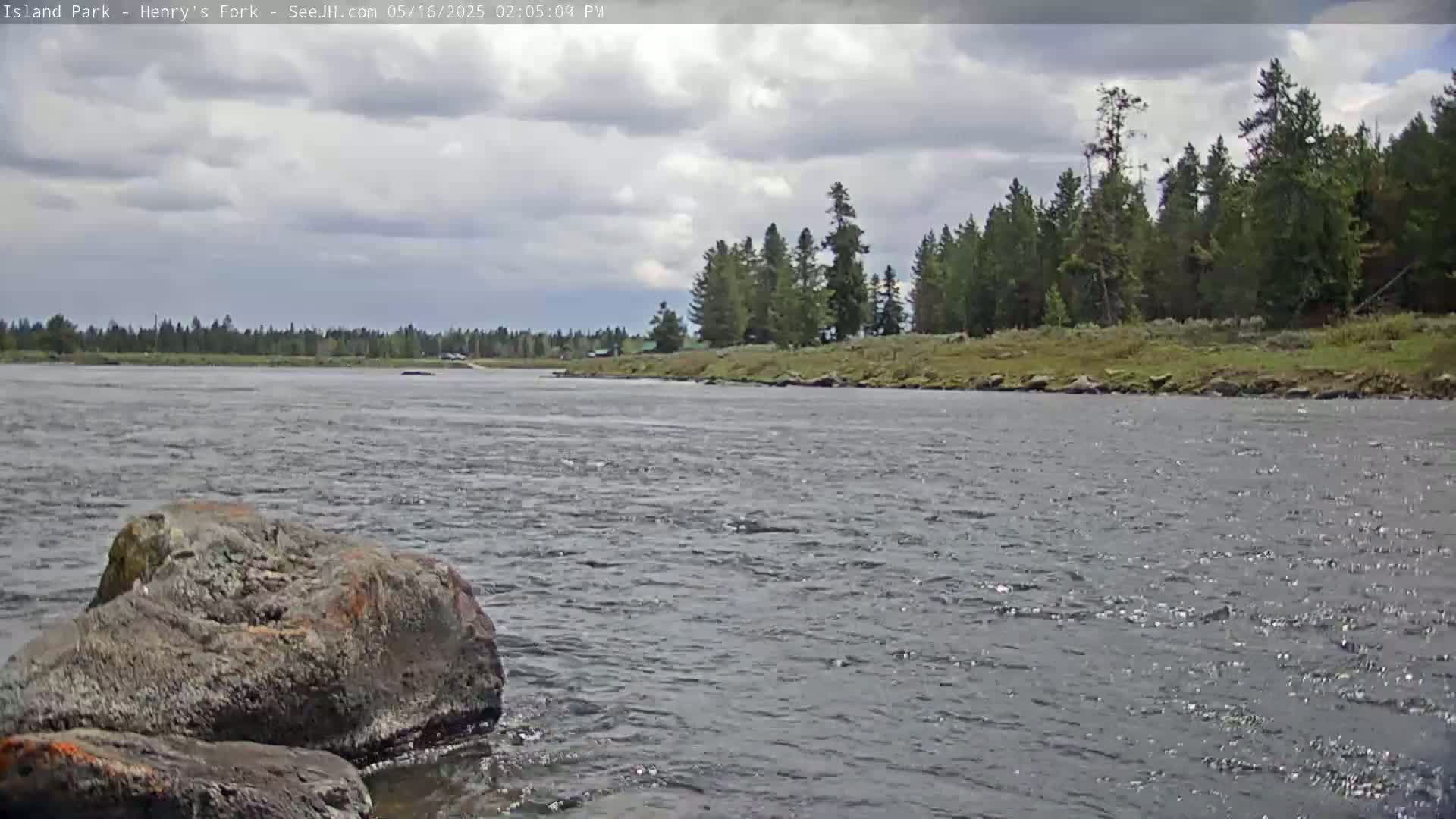 A partly cloudy sky overlooks a wide river with a rocky shoreline lined with coniferous trees.