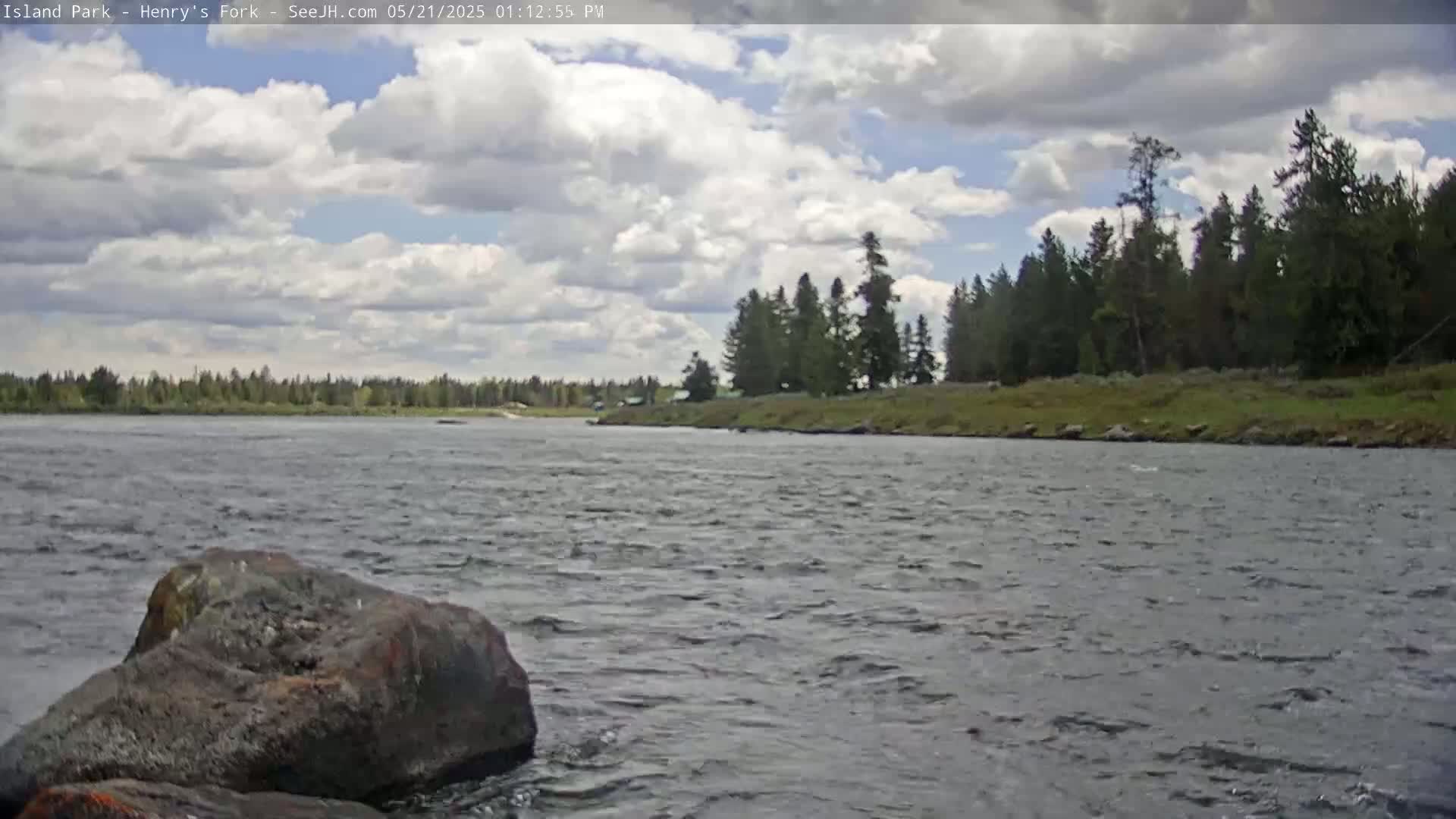 A partly cloudy sky overlooks a wide river with a rocky bank lined with evergreen trees.