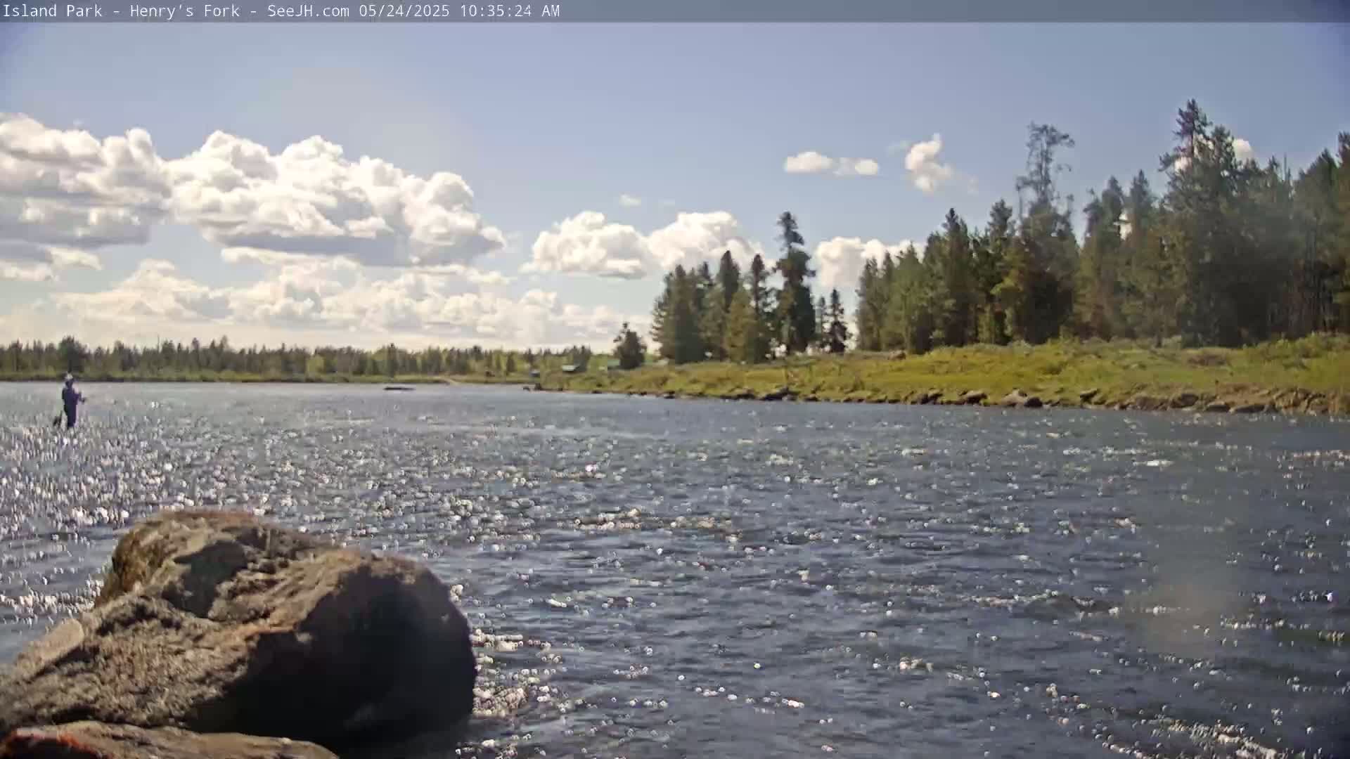 A person fly fishes in a swiftly flowing river under a partly cloudy sky.