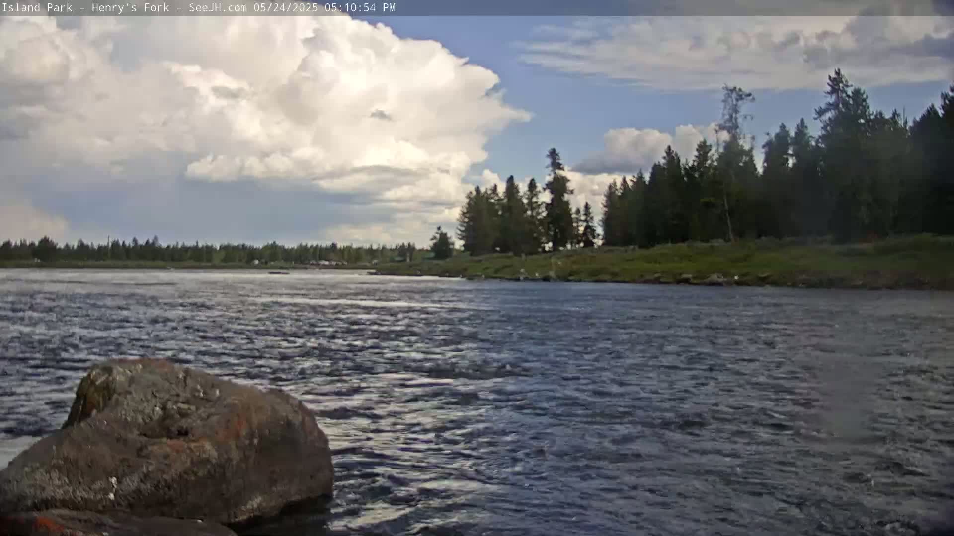 A partly cloudy sky hovers over a wide river flowing past a rocky shore and a line of evergreen trees.