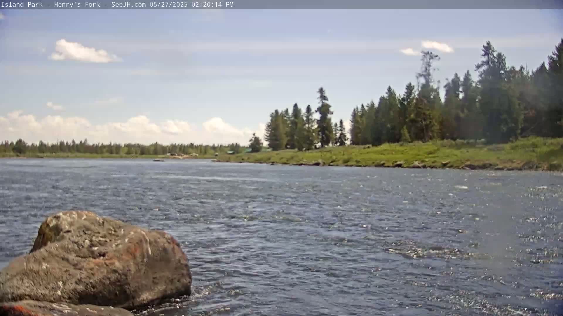 A partly cloudy sky overlooks a river flowing past a rocky bank and a line of evergreen trees.
