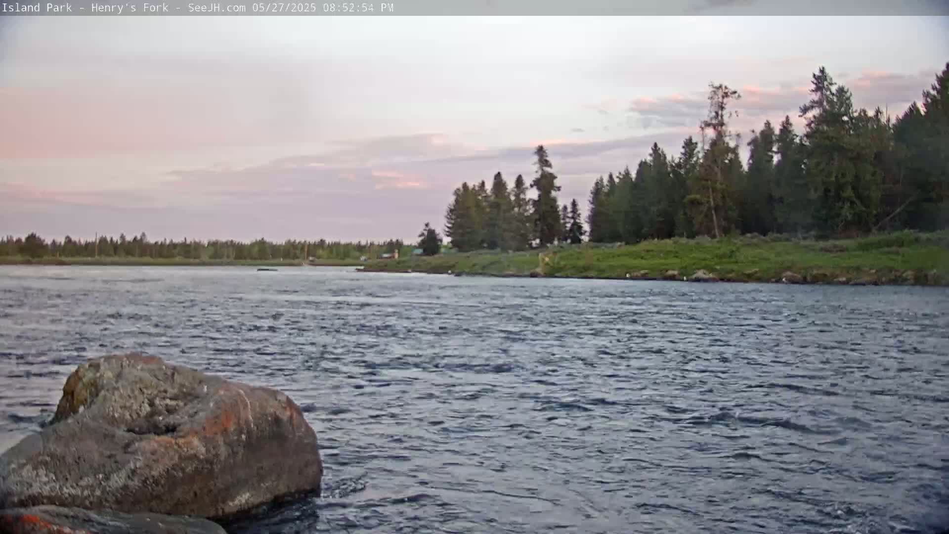 A large rock sits in the foreground of a flowing river under a partly cloudy, pinkish-hued sky, with a line of trees visible in the distance.