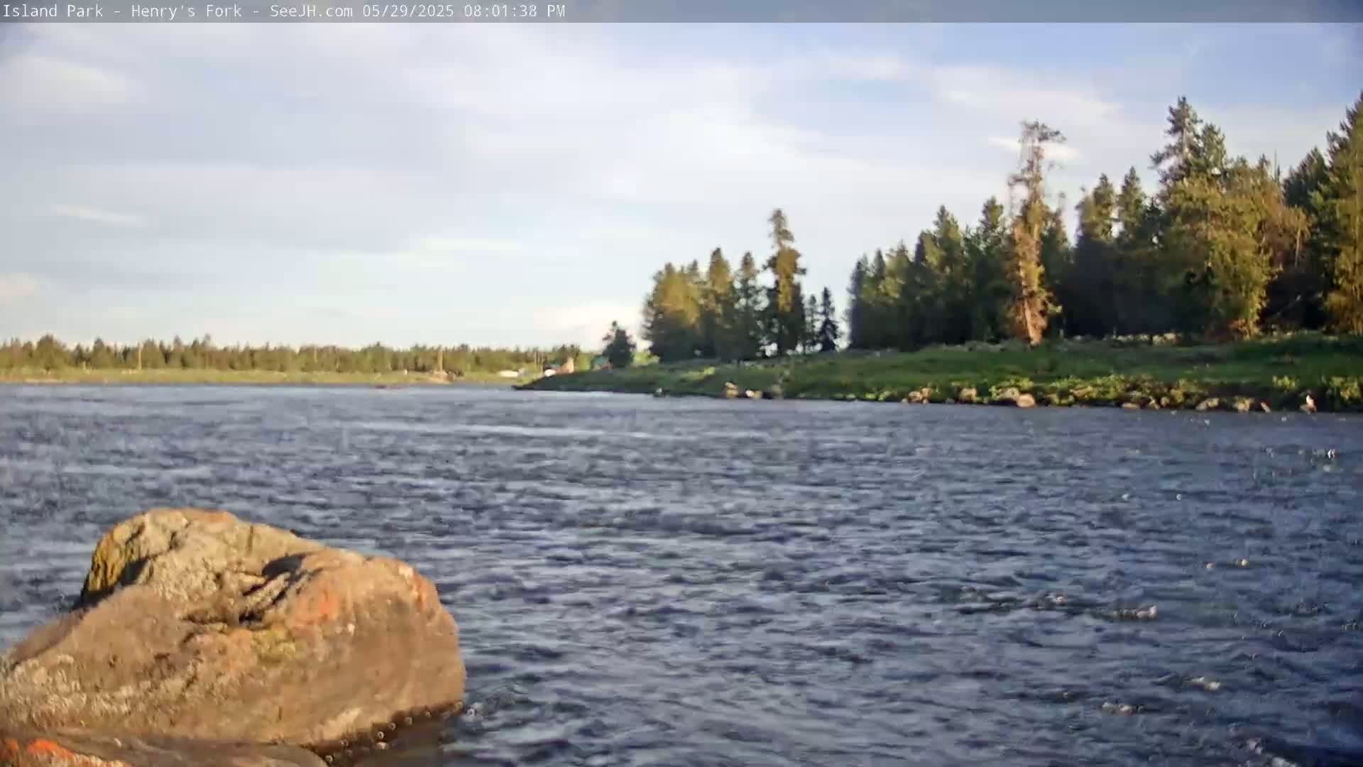 A rocky river flows under a partly cloudy sky past a treeline on a sunny afternoon.