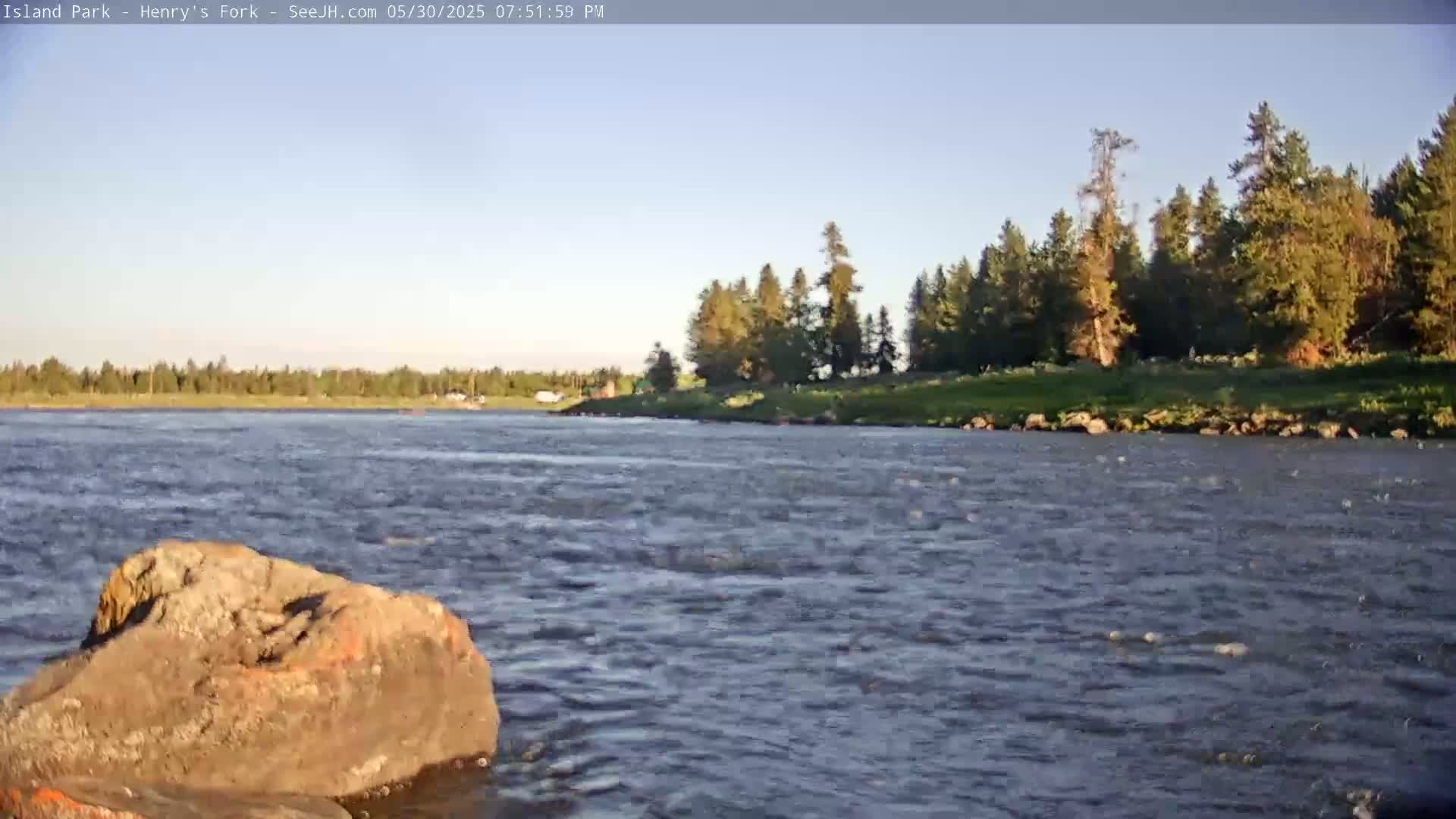 A large, brown rock sits in the foreground of a gently flowing river under a clear, light blue sky, with a line of trees visible on the far bank.