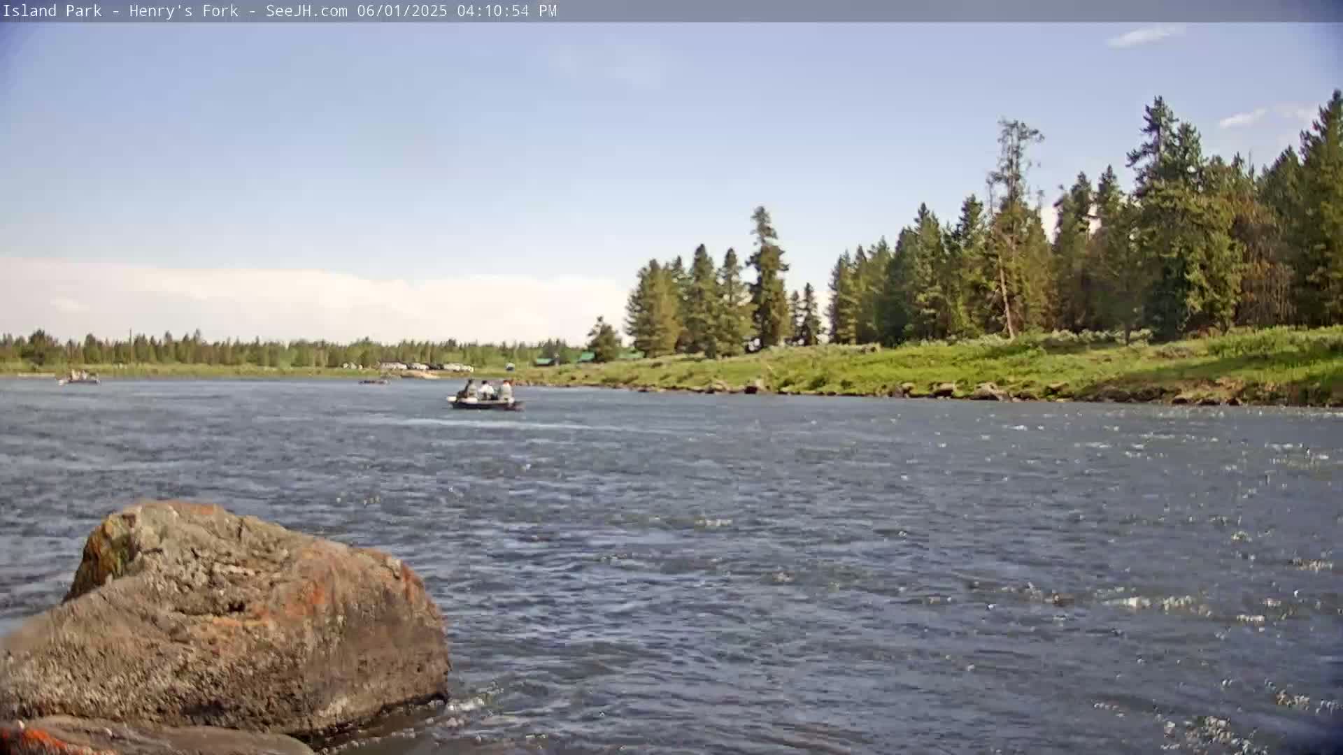 A mostly sunny day shows a wide river with a few small boats and a rocky shoreline lined with green trees.