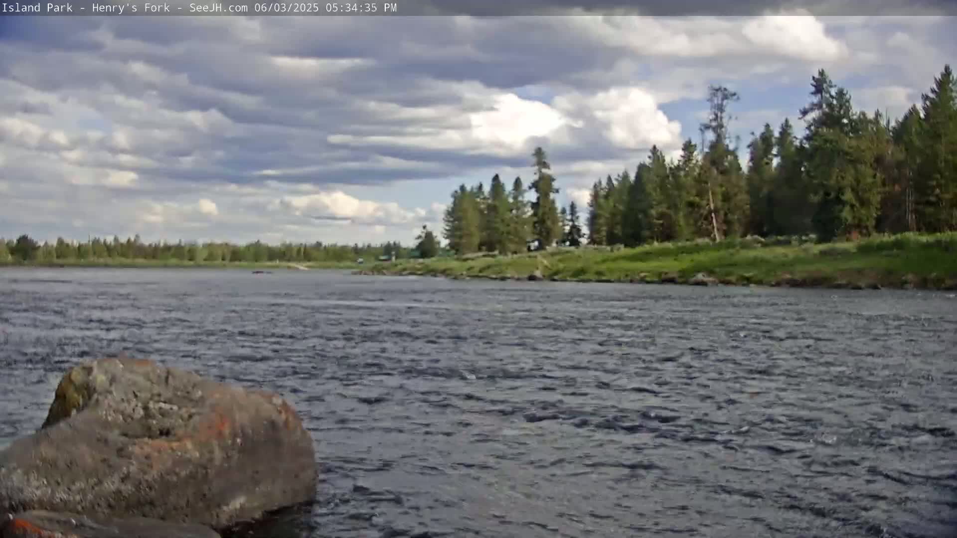 A partly cloudy sky overlooks a river flowing past a rocky bank and a line of evergreen trees under a partly cloudy sky.