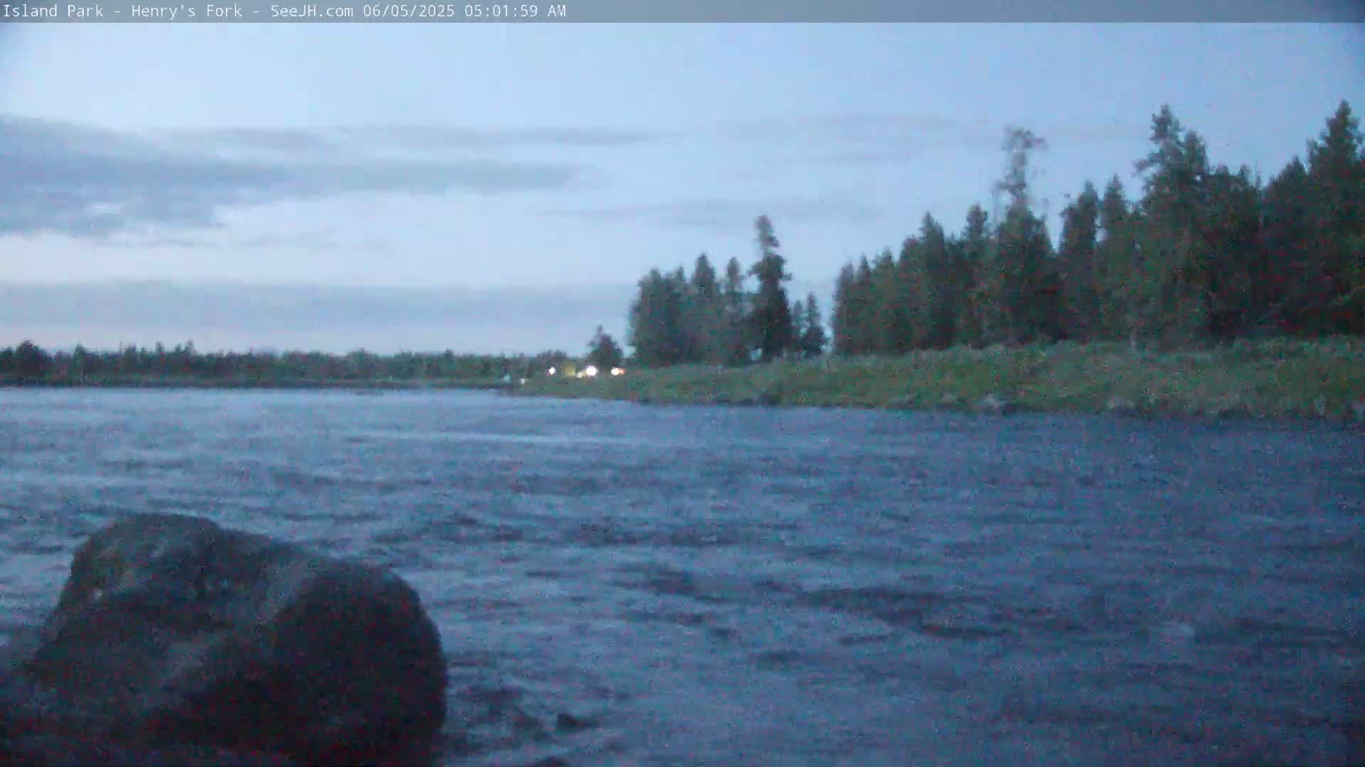 A dark, choppy river flows under a twilight sky, past a line of evergreens on the far bank.