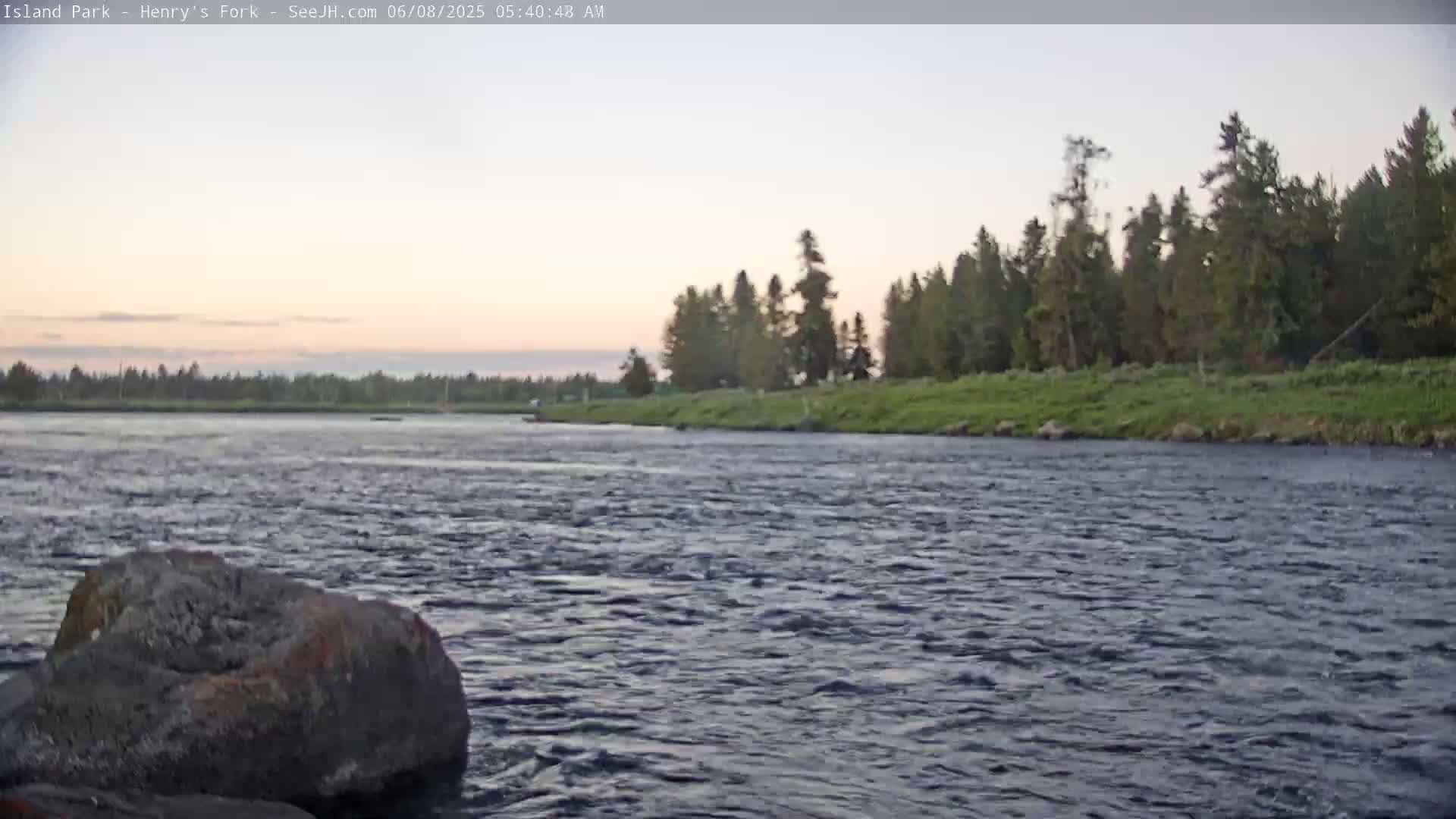 A rocky river flows past a verdant shoreline lined with trees under a calm, dusky sky.